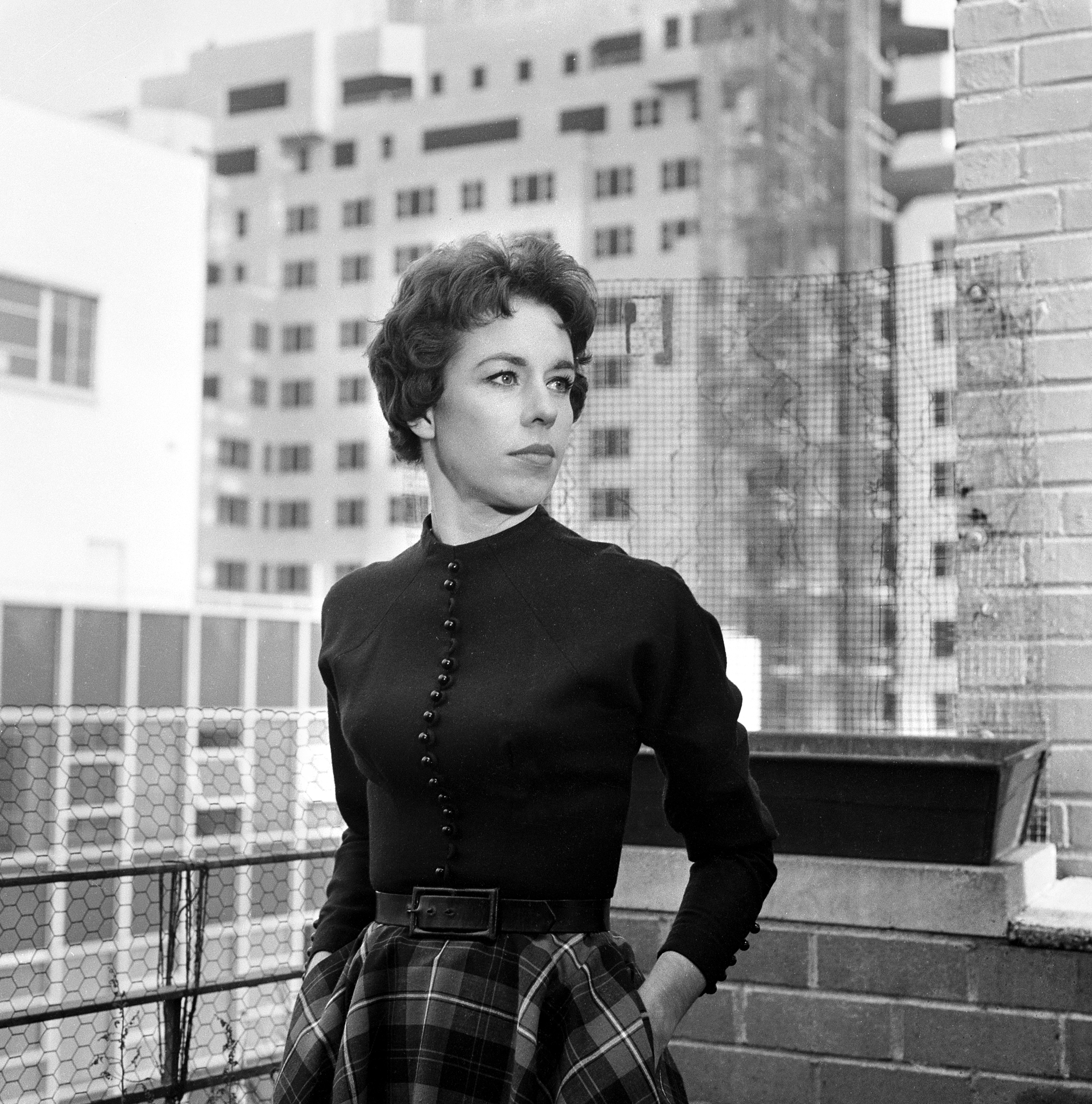 Standing on a rooftop terrace, she wears a long-sleeved, buttoned black top tucked into a high-waisted plaid skirt, cinched with a belt. Her hands rest in her pockets as she looks off to the side, with city buildings visible behind her.