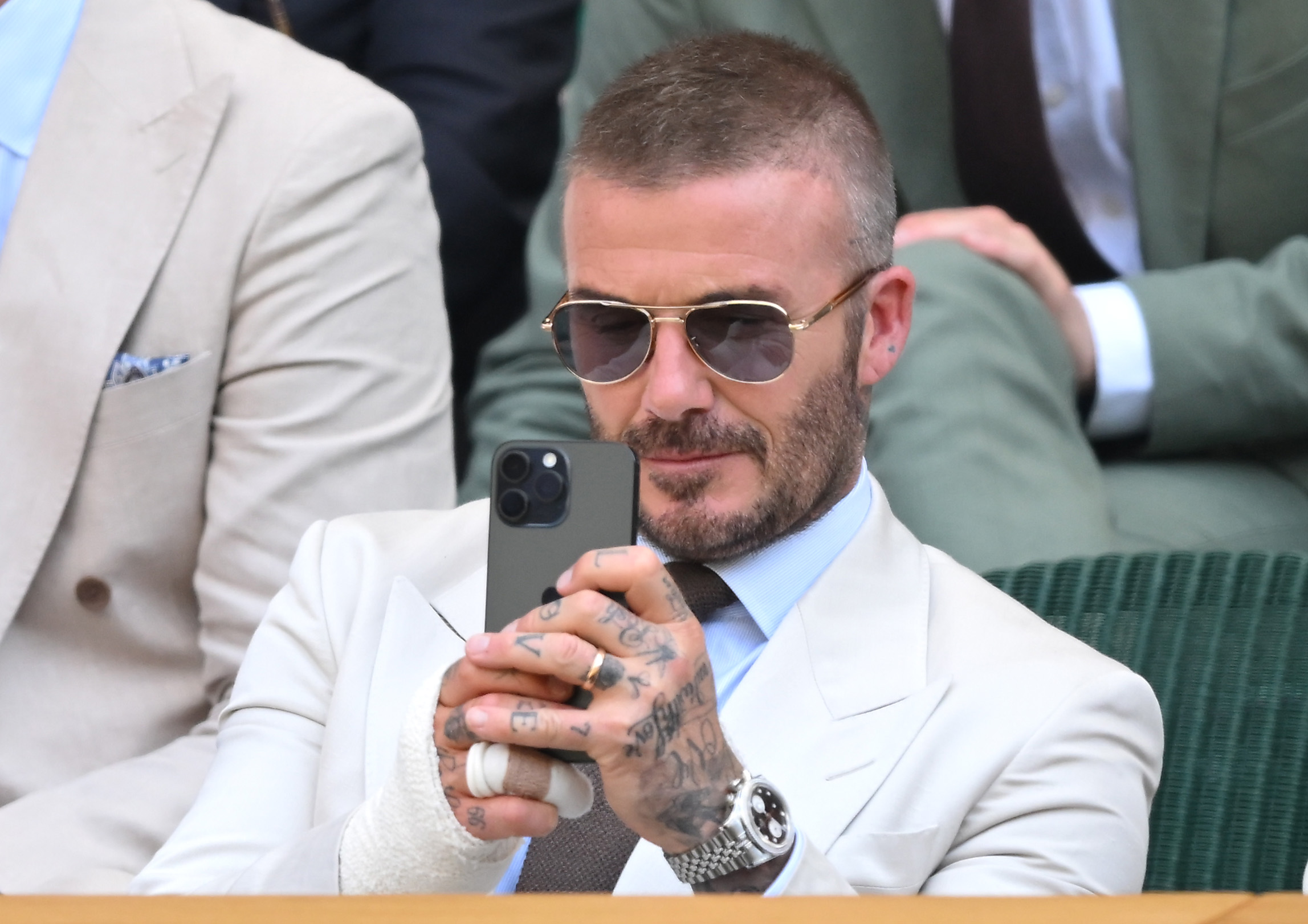 David Beckham uses his phone while seated courtside at Wimbledon. His sunglasses and tailored suit add to his polished look, while his bandaged hand remains clearly visible.