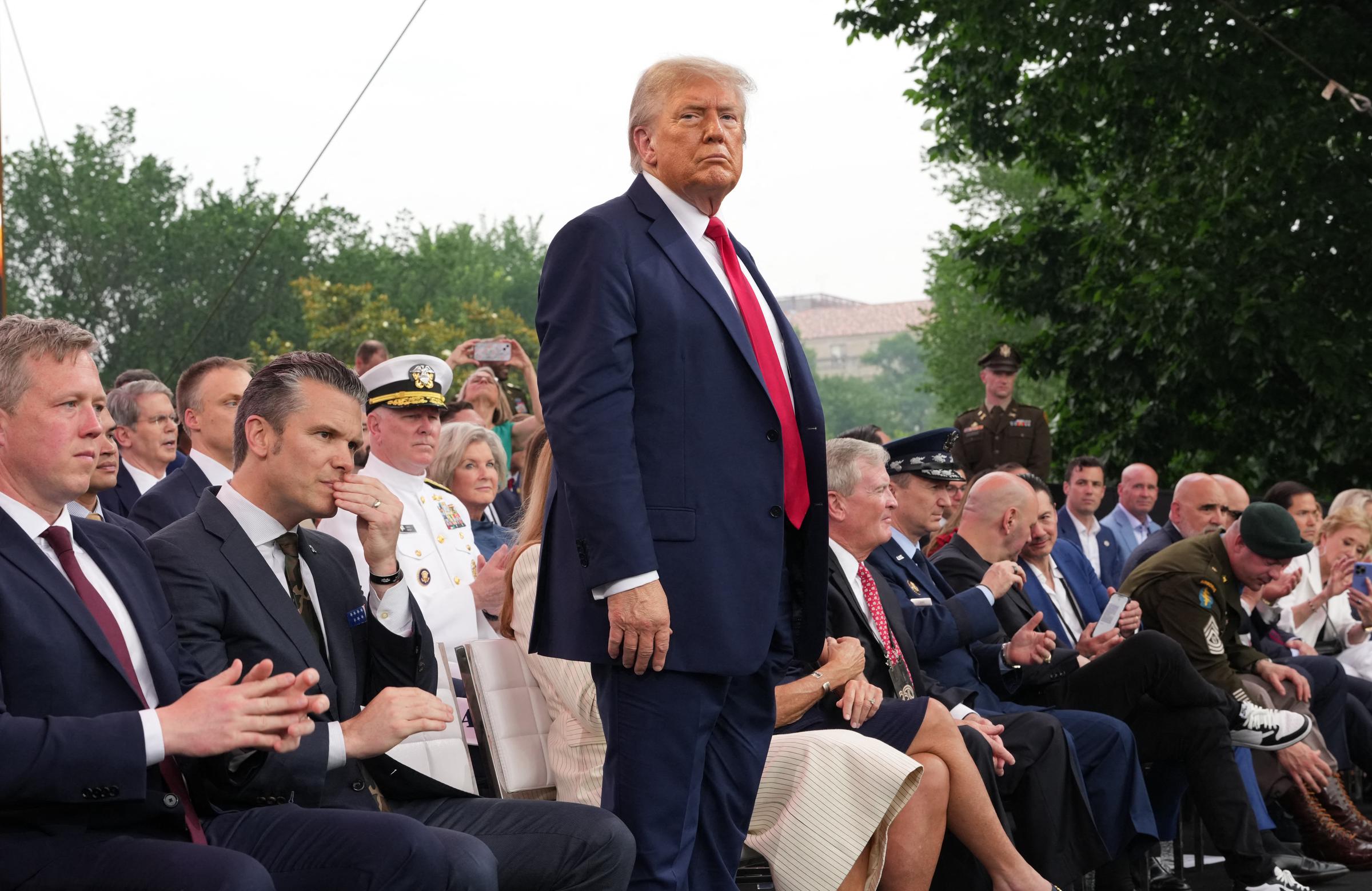 Donald Trump stands among seated dignitaries, military leaders, and guests on stage during the U.S. Army’s 250th anniversary parade on June 14, 2025. His upright stance, with Melania seated nearby, draws attention amid the formal, attentive audience.