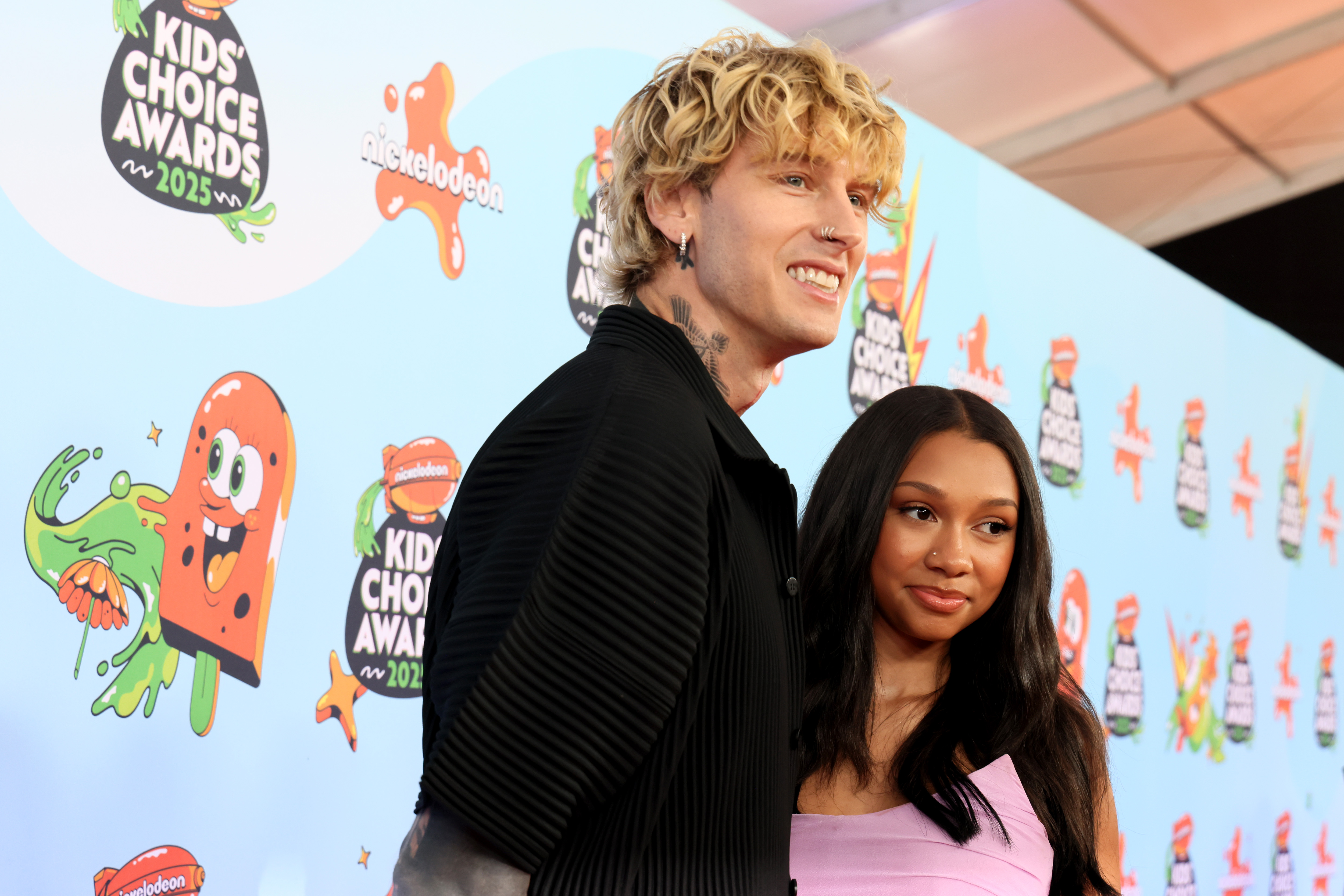 MGK and Casie Baker arrive together at the 2025 Nickelodeon Kids' Choice Awards in Santa Monica on June 21, posing with relaxed smiles against the event's bold, animated backdrop.