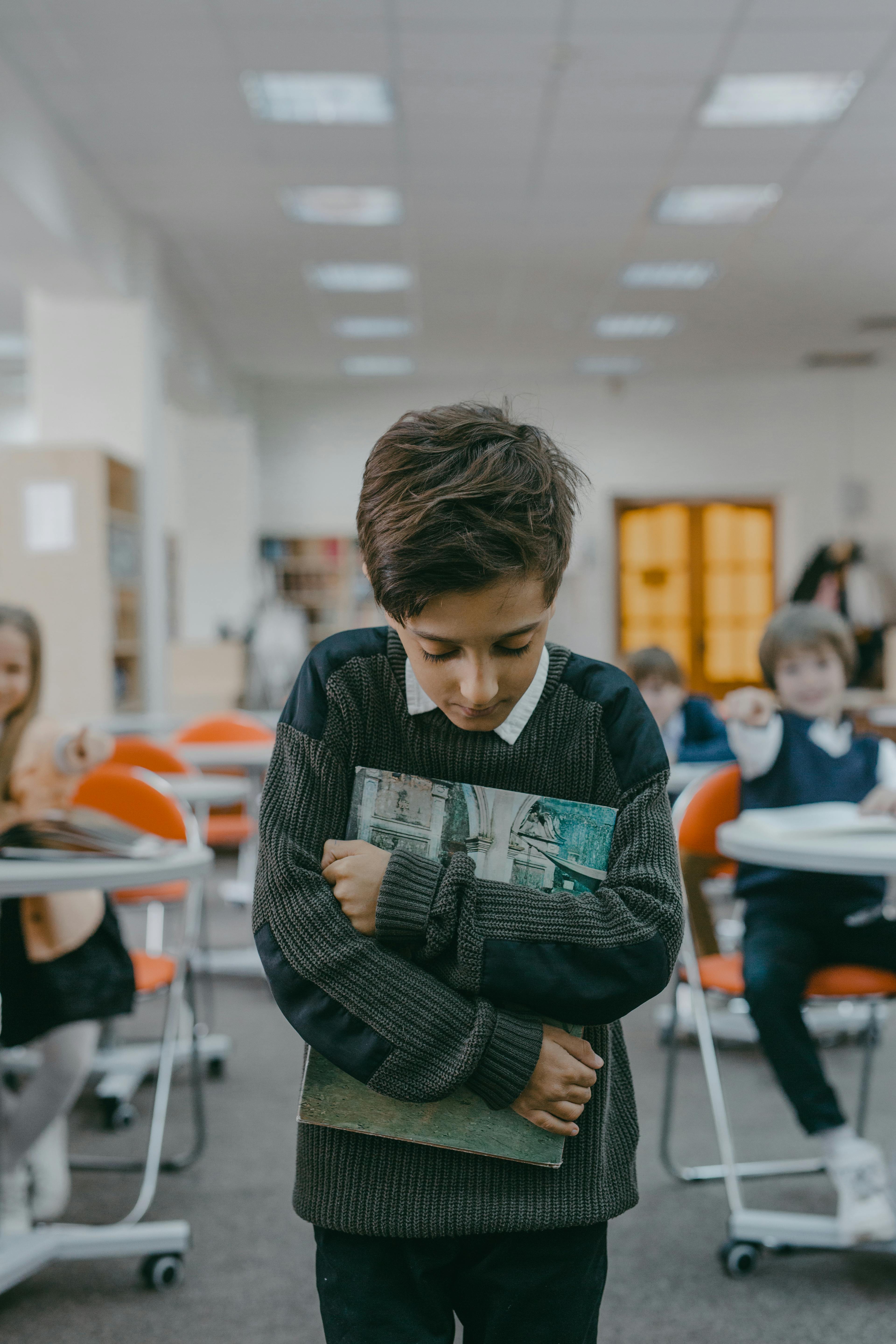 A student clutches a book tightly while others in the background appear to whisper and point, suggesting exclusion and distress in the classroom. | Source: Pexels