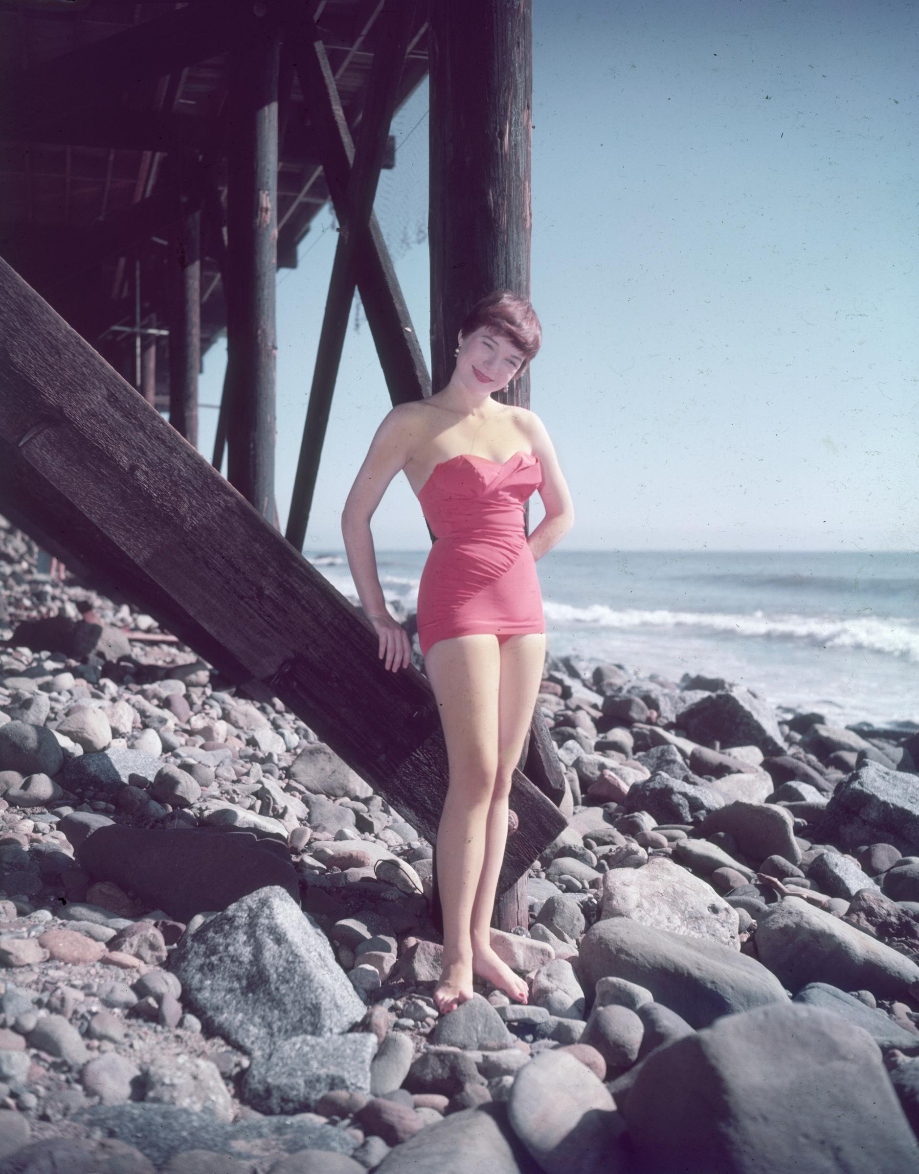 Shirley MacLaine in a pink strapless swimsuit standing on rocks beneath a wooden pier, circa 1955.