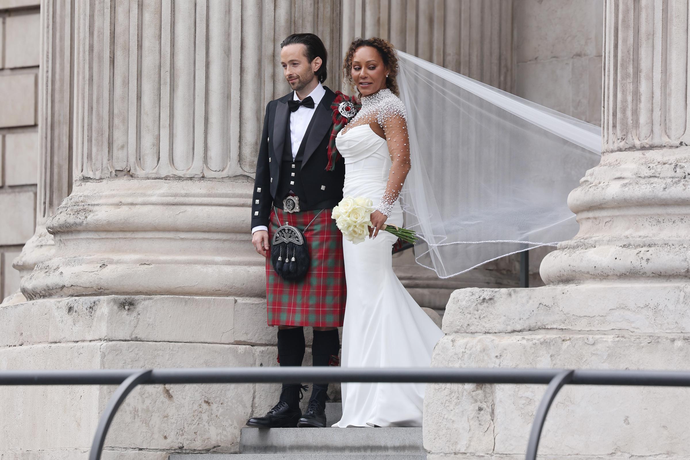 In a full-length image, Mel B and Rory McPhee stand together on the steps of St. Paul’s Cathedral. The iconic structure towers behind them as they pose hand in hand, their wedding attire crisp against the historic stone. The photo captures the grandeur of the day and the quiet joy between the couple.