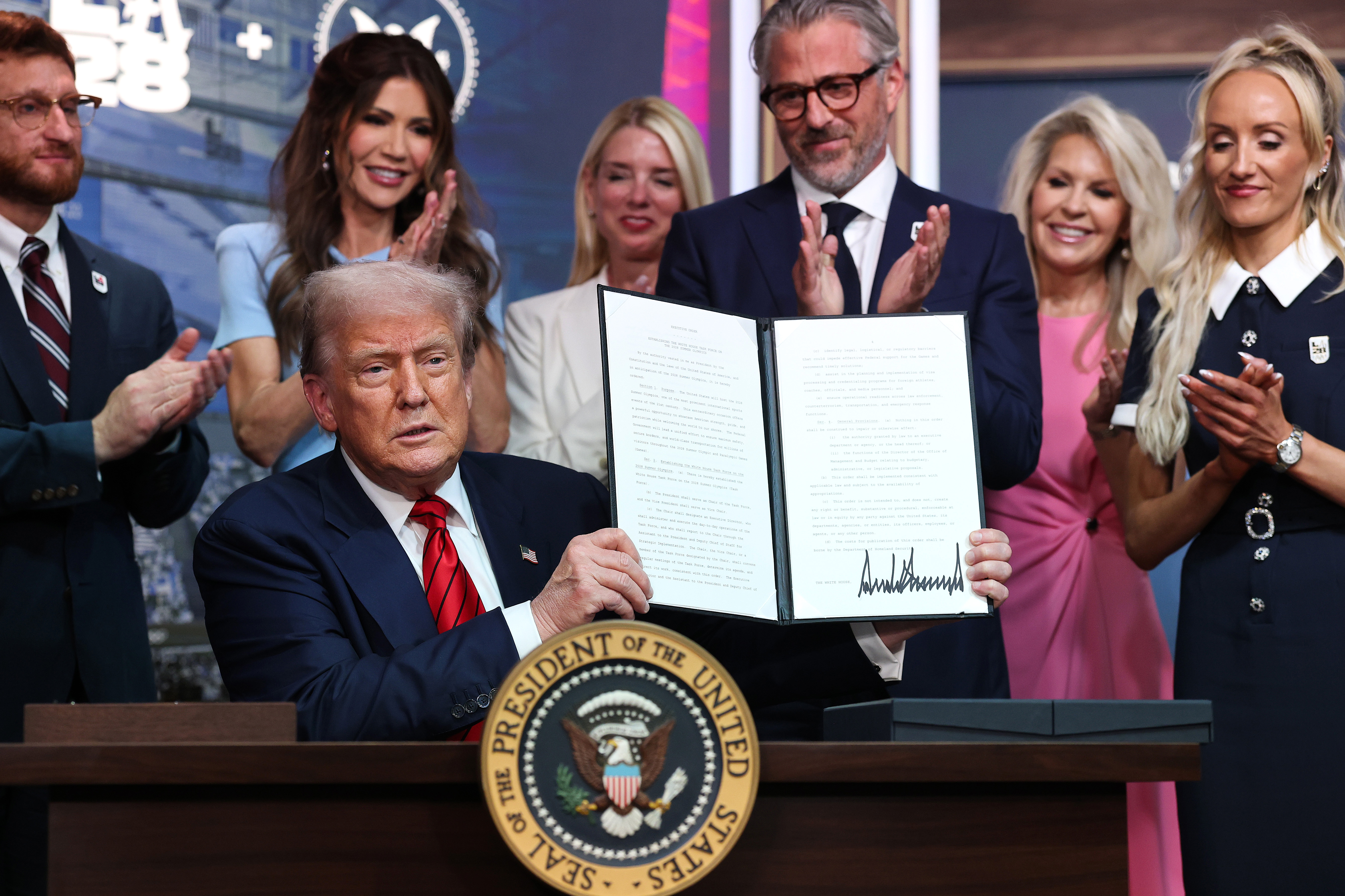 Holding the signed executive order high, Trump presents it to the audience at the White House's South Court Auditorium while officials and guests cheer.