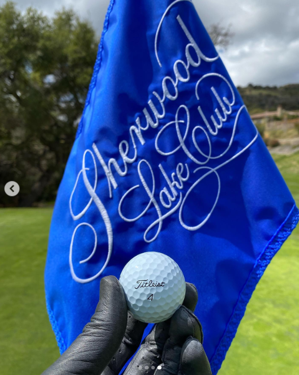 Brandon Thomas Lee holds a golf ball in front of a blue flag printed with the Sherwood Country Club logo, posted in March 2021 | Source: Instagram/brandonthomaslee