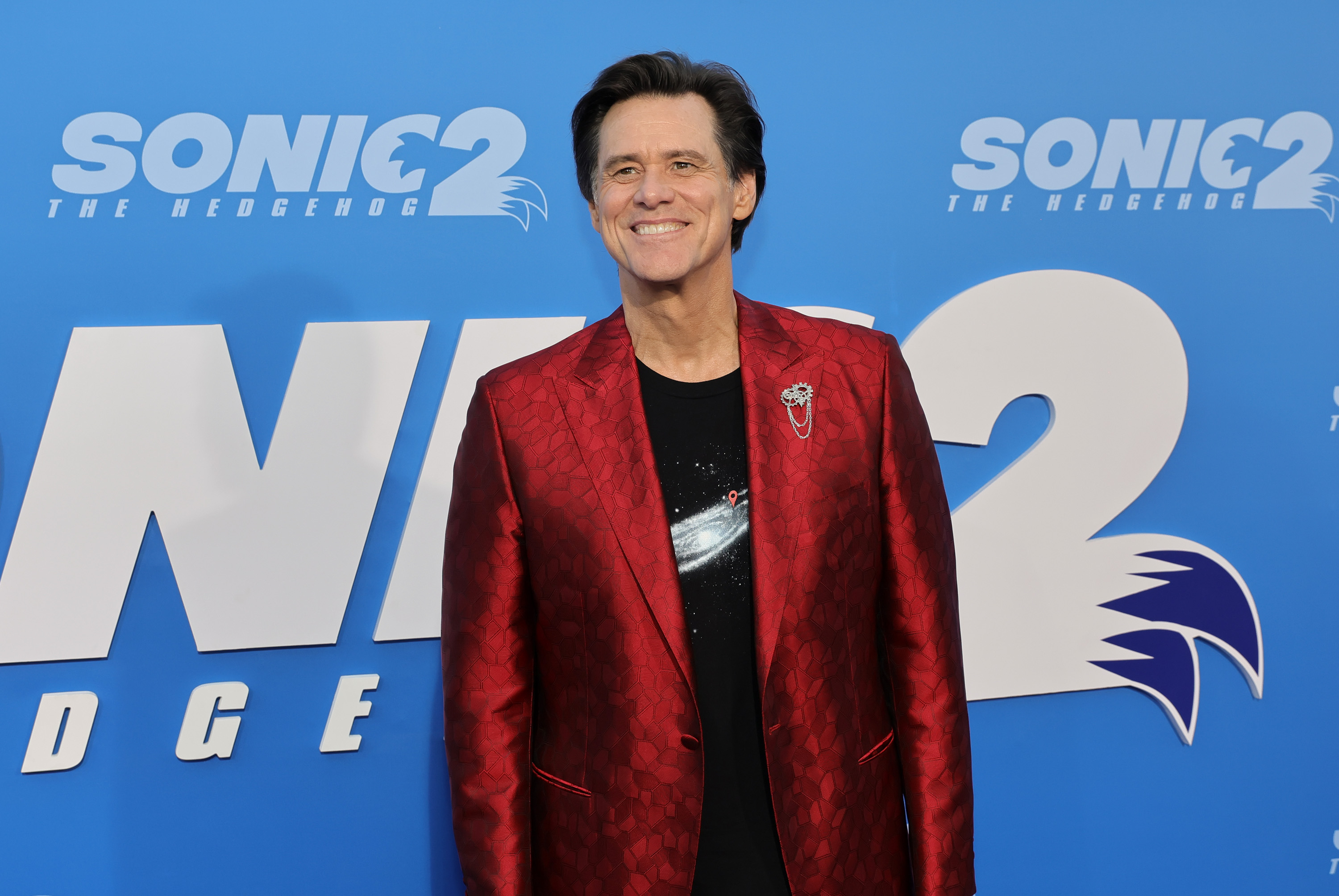 The photo shows Jim Carrey attending the Los Angeles premiere screening of "Sonic the Hedgehog 2" at the Regency Village Theater. He is dressed stylishly in a red patterned blazer over a black shirt featuring a galaxy design. Smiling confidently against the blue event backdrop, he exudes charisma and star power.