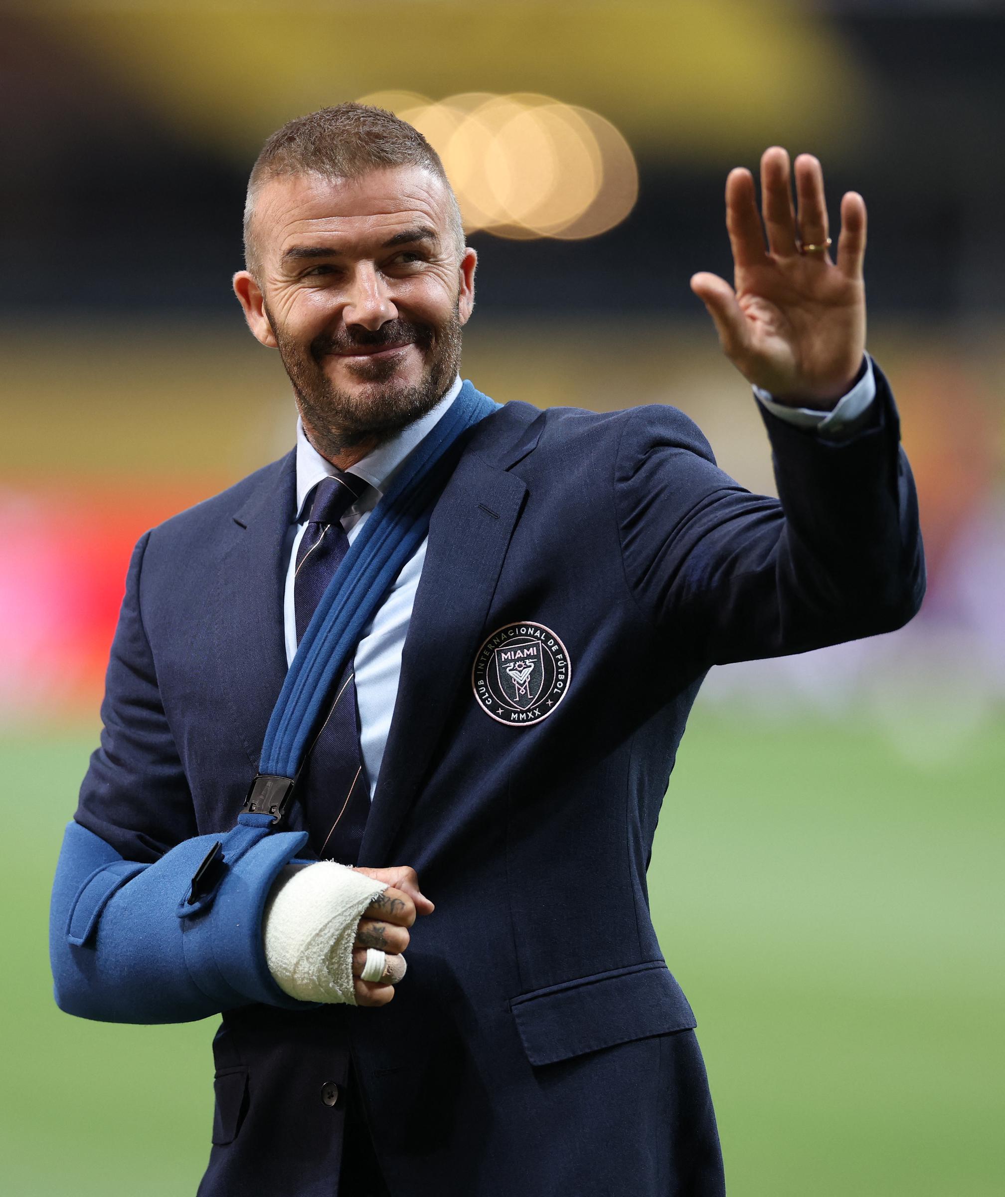 David Beckham waves to fans at Mercedes-Benz Stadium on June 29. Wearing a navy suit and blue arm sling, his right hand is visibly bandaged, but his confident smile stands out.