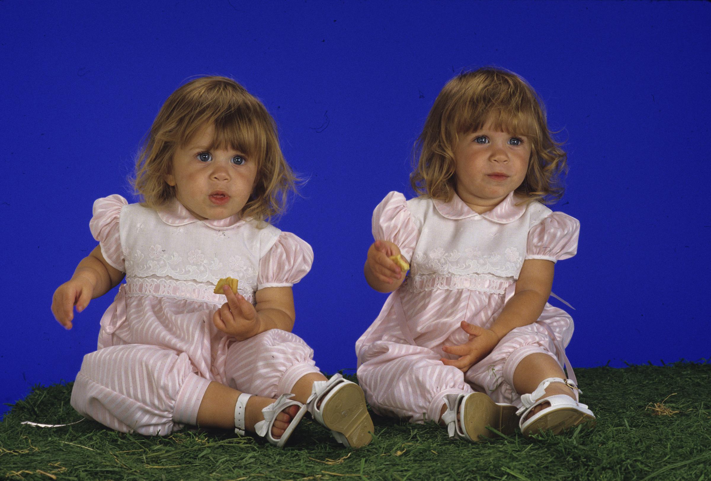 Mary-Kate Olsen and Ashley Olsen sit side by side as toddlers in matching pale pink outfits. They hold snacks and look forward with curious, wide-eyed expressions.