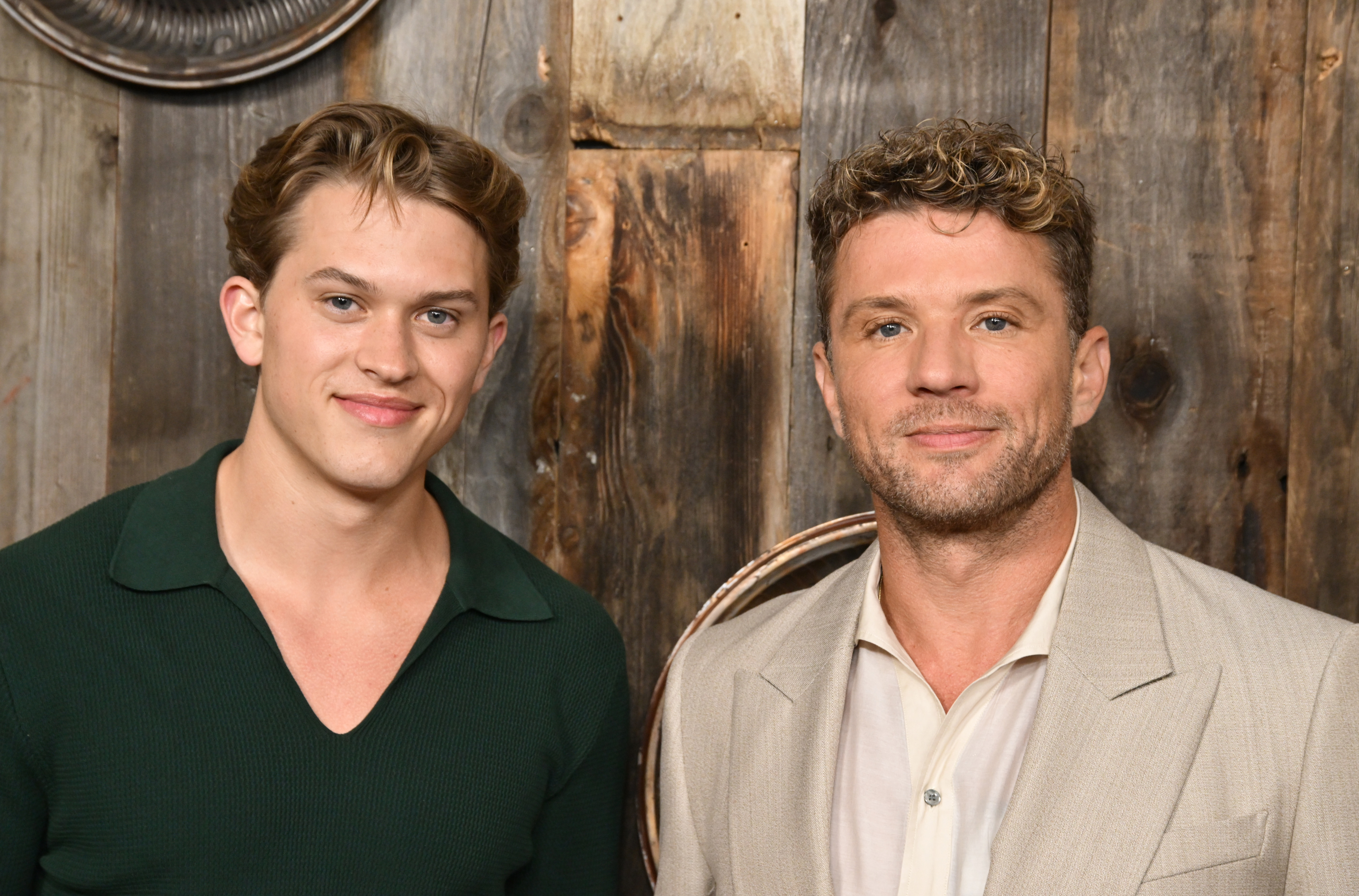 The father-son duo flashes friendly smiles as they face the cameras at the "Motorheads" red carpet, with Deacon in a sharp forest green top and Ryan in a laid-back sand-colored suit, posing against the wood-paneled photo wall.