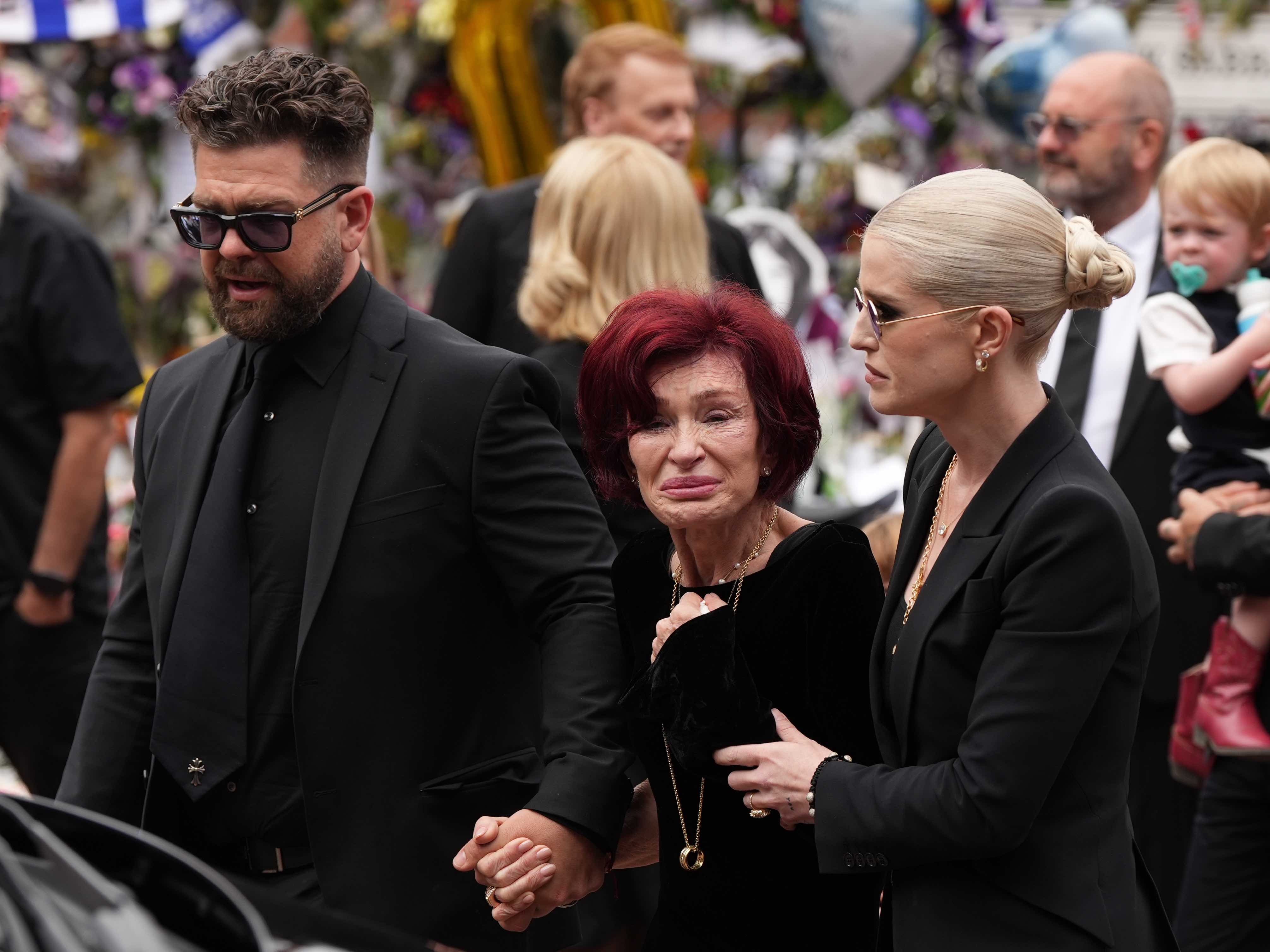 Sharon Osbourne, wife of Ozzy Osbourne, attends Ozzy's funeral as his funeral cortege arrives at the Black Sabbath Bridge bench. Sharon clutches the hands of her children, Jack and Kelly, during this heart breaking moment.