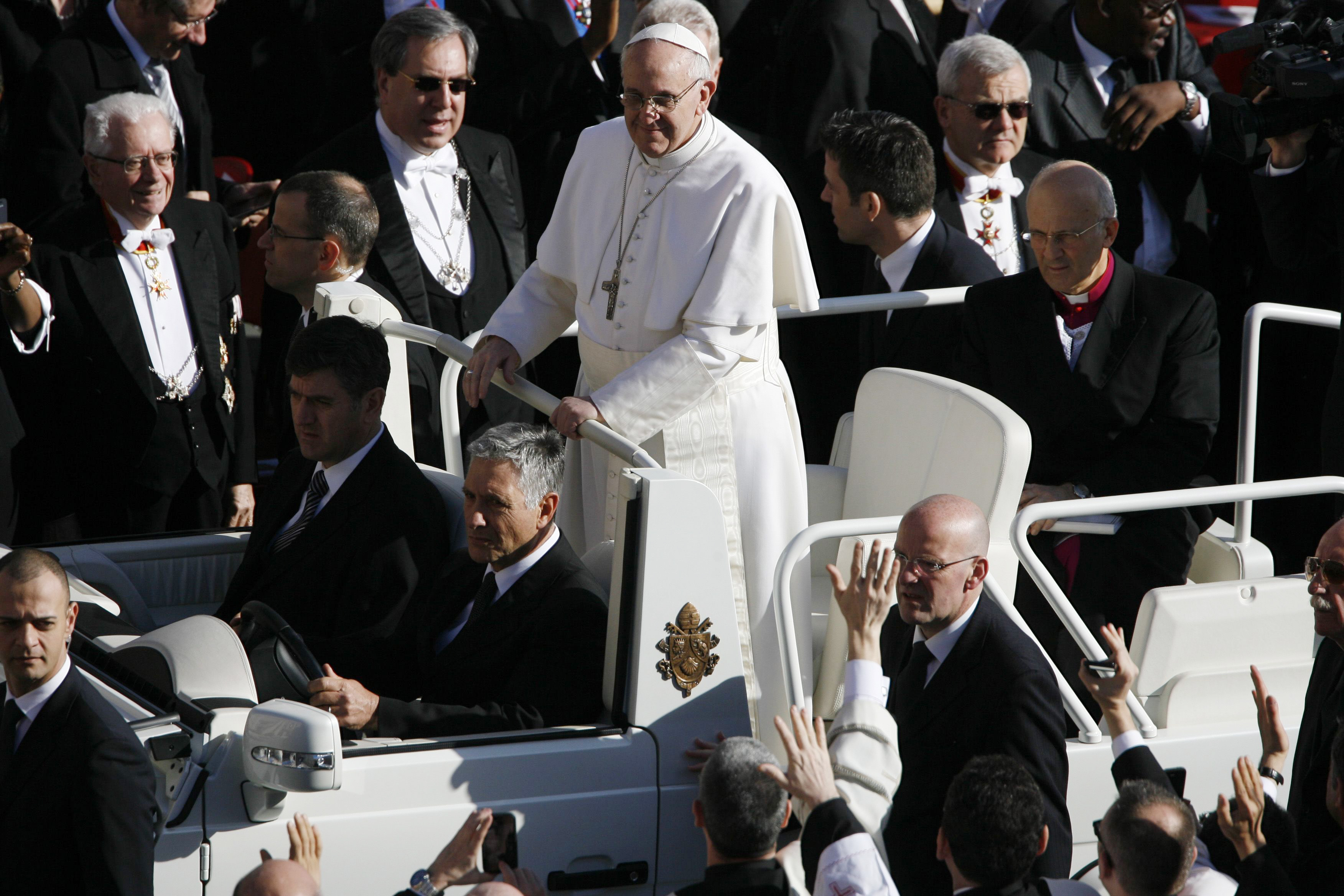 Pope Francis stands in the popemobile surrounded by a sea of dignitaries and waving hands, dressed in full white with a silver cross draped over his chest