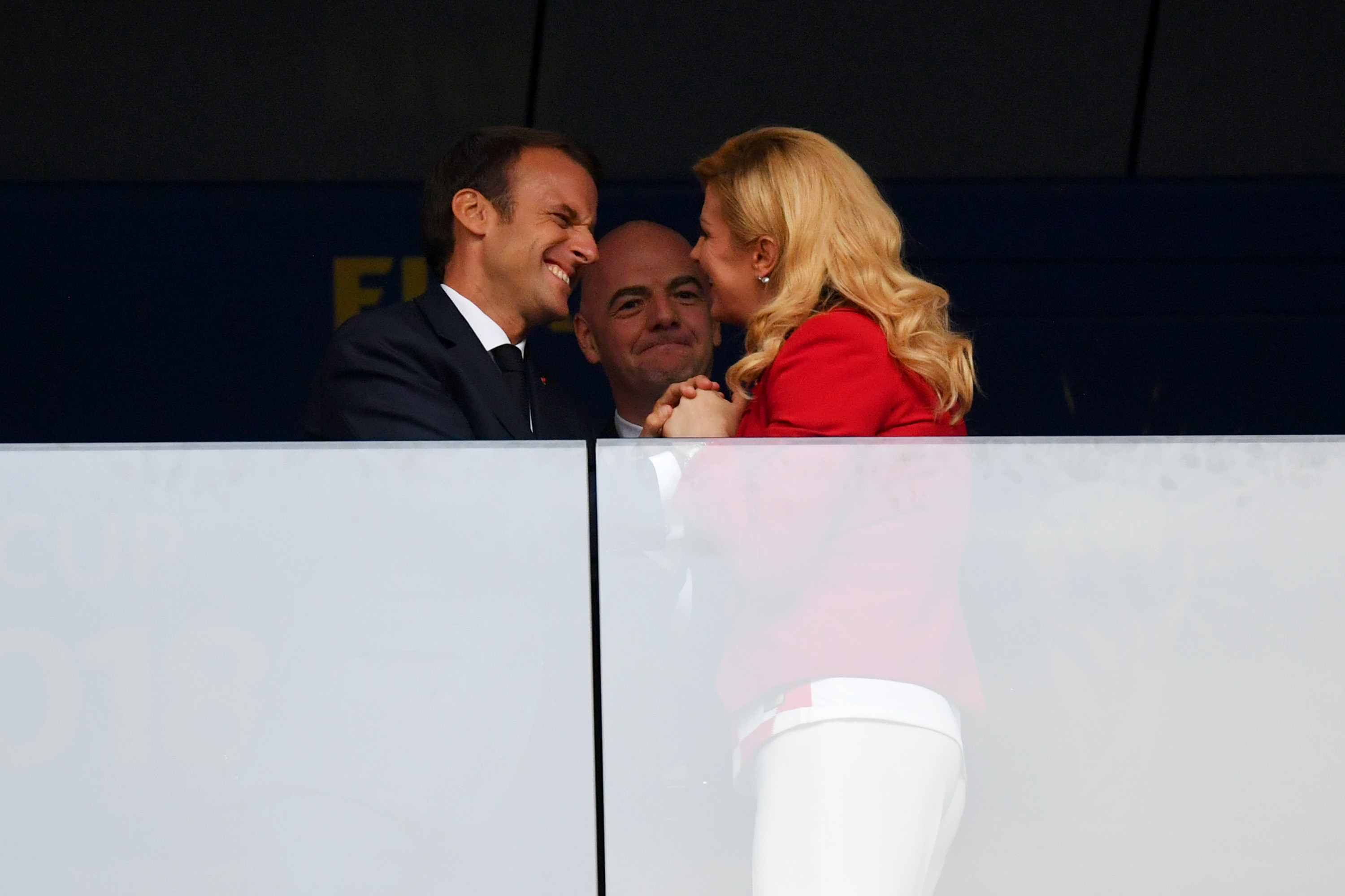 Emmanuel Macron and Kolinda Grabar-Kitarović share a cheerful moment as they lean in close, smiling and maintaining hand contact while Gianni Infantino watches behind them.