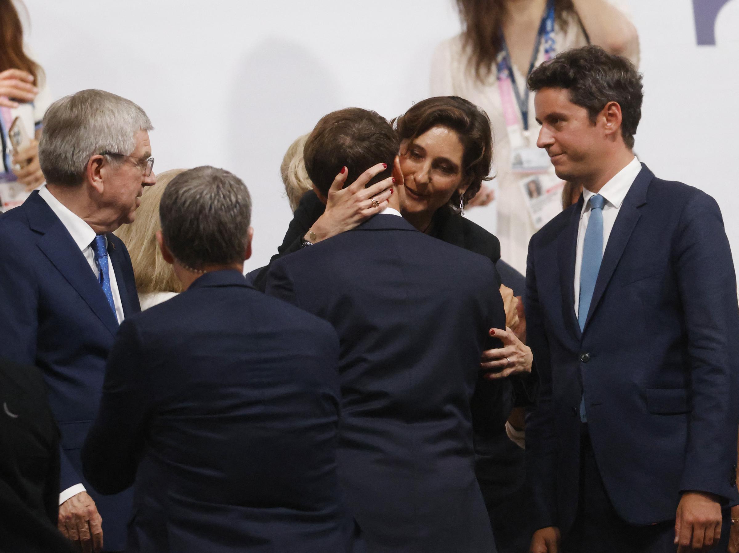 In another frame, Amélie Oudéa-Castéra maintains her hold around Emmanuel Macron’s shoulders while pressing her cheek against his. The surrounding officials remain engaged in conversation, with one man glancing toward the pair.