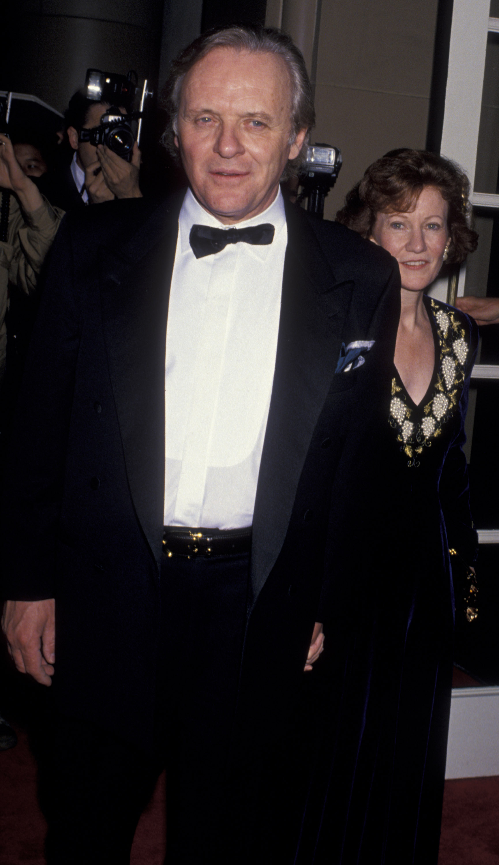Anthony Hopkins and Lyn Davis grace the red carpet at the Fifth Annual BAFTA Awards, radiating timeless elegance. Hopkins, in a classic black tuxedo and bow tie, stands poised under the flash of cameras. Beside him, Davis exudes quiet sophistication, her embroidered gown adding a touch of refined glamor to the evening.
