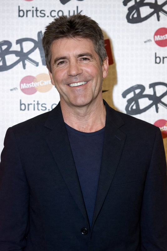 Dressed in a navy blazer over a matching crew-neck top, he grins broadly at what appears to be a Brit Awards media event, grey beginning to show at the temples. | Source: ChatGPT