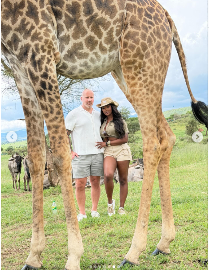 Framed beneath towering giraffe legs, Ashly Robinson and Joe McCann stand close in the open savannah. | Source: Instagram/ashleejenae