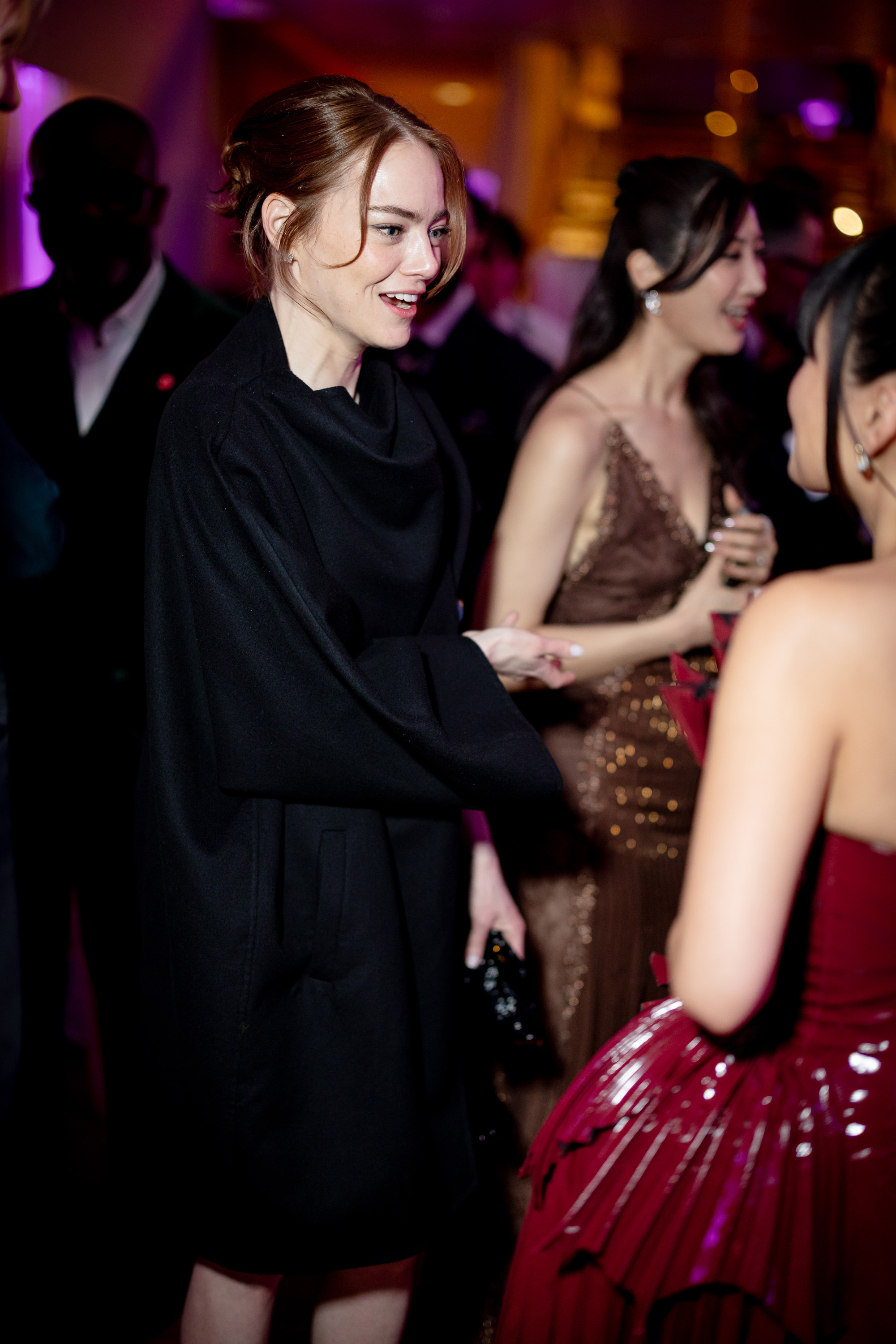 Emma Stone chats with Rei Ami during the BAFTA Dinner, wearing a relaxed black mini dress with a draped cape as the pair mingle in the softly lit reception space.