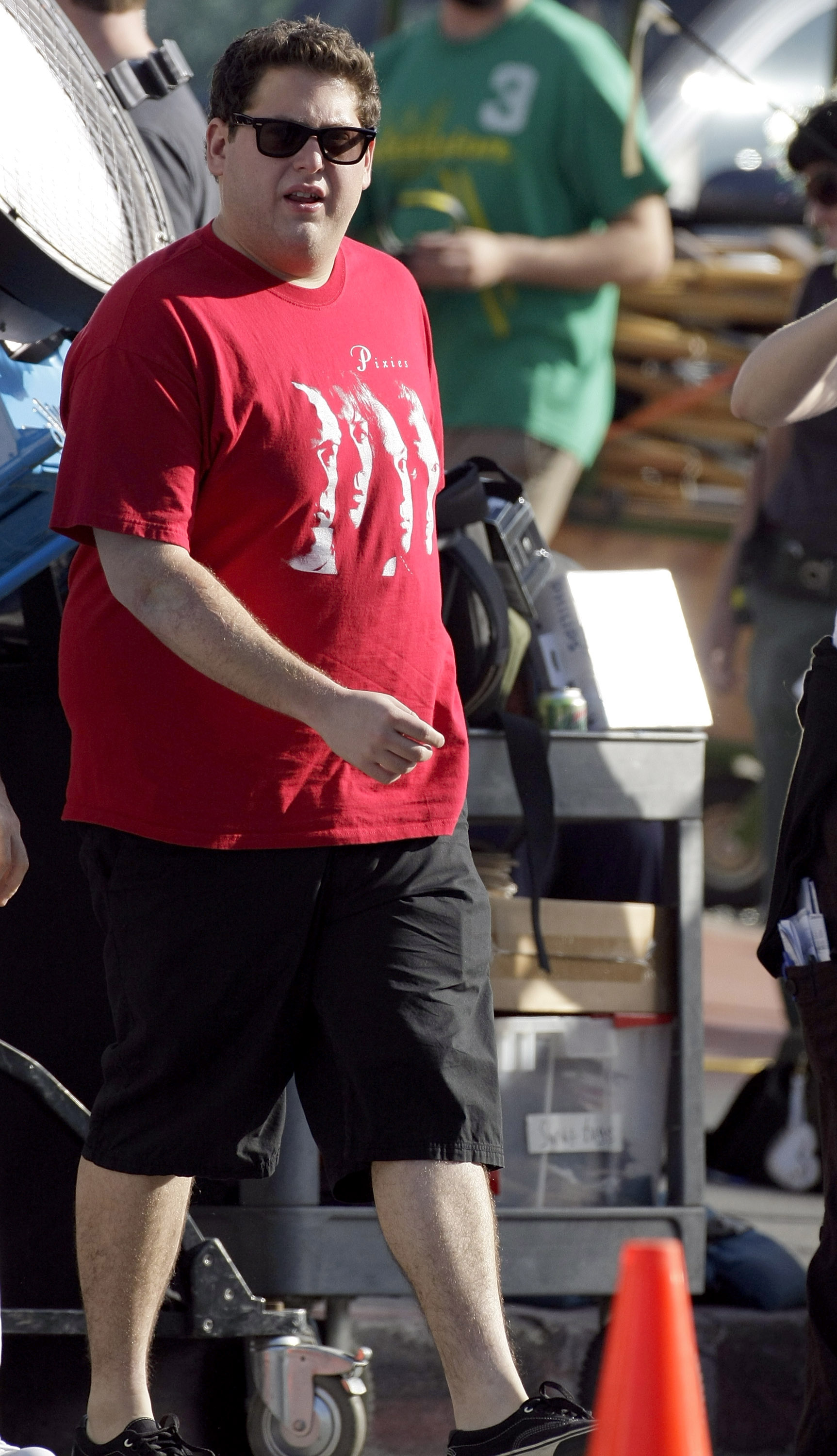 Jonah Hill is spotted on set wearing a red Pixies band tee, black shorts, and dark sunglasses. Walking between equipment carts and crew members, he keeps it casual in sneakers while filming outdoors.