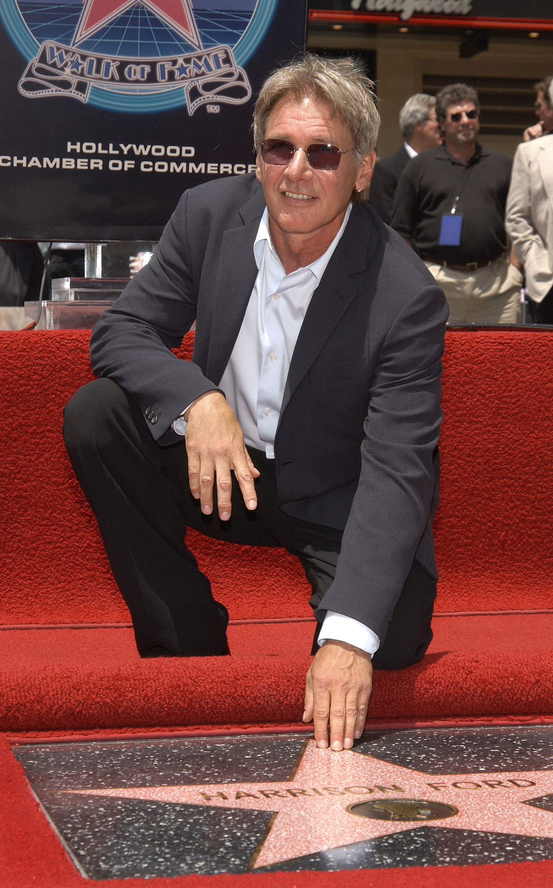 Kneeling on the red carpet, Ford smiles as he places his hand on his newly unveiled star during his Hollywood Walk of Fame ceremony. Dressed in a textured charcoal blazer, a crisp white button-down, and tinted sunglasses, the actor celebrated this major career milestone in Los Angeles. | Source: Getty Images
