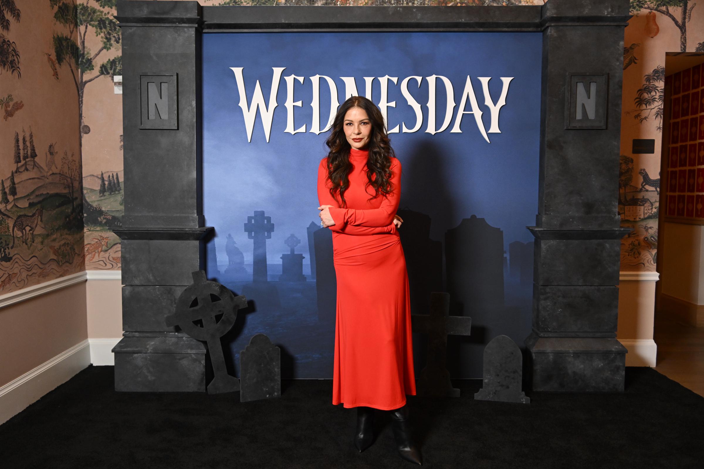 Wednesday SAG Nom Comm, New York, November 11, 2025 — Zeta-Jones poses arms-folded in a long-sleeved red turtleneck maxi dress and black knee-high boots in front of a gothic "Wednesday" Netflix backdrop featuring a graveyard set, her dark wavy hair loose over her shoulders.