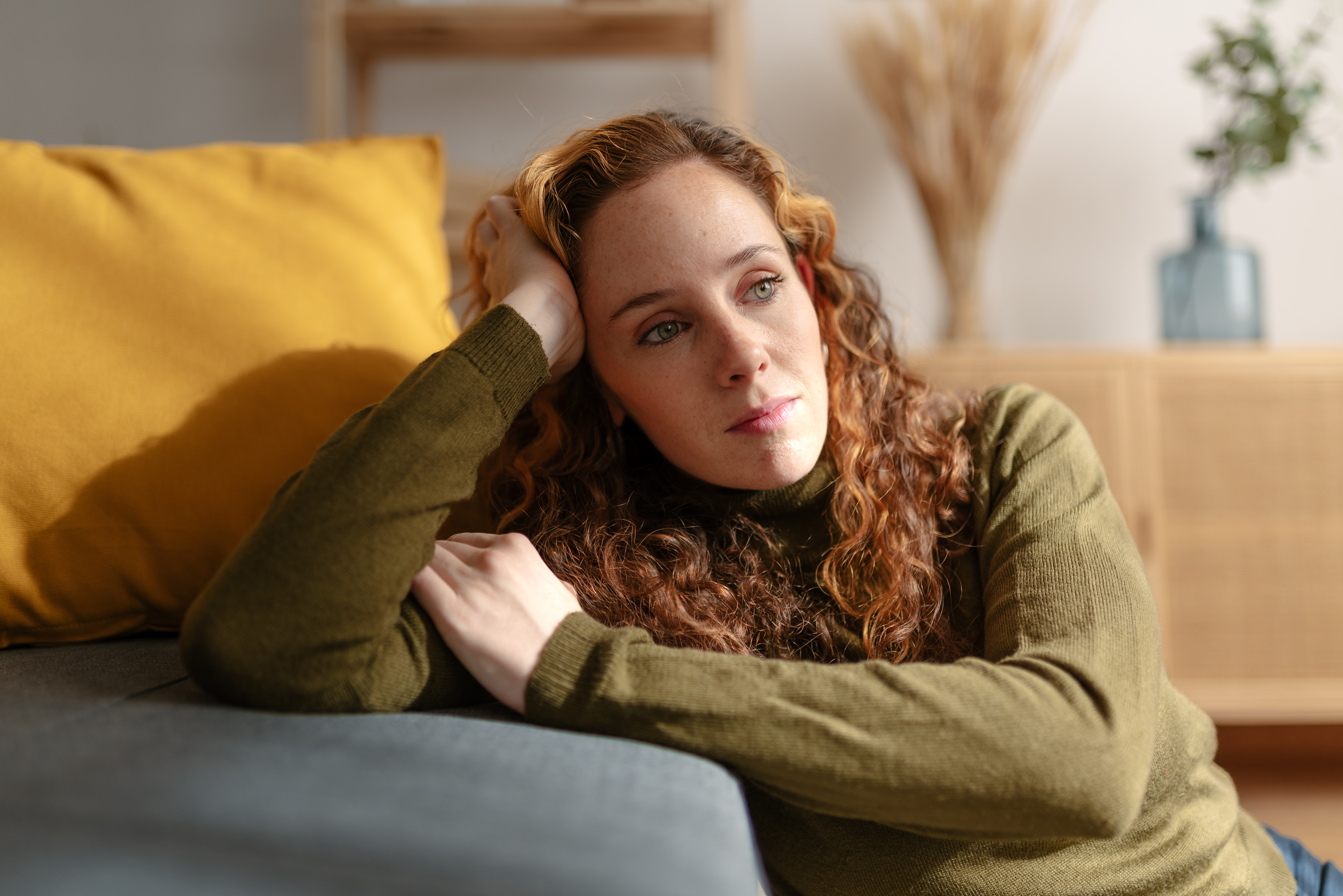 Portrait of a young woman with long curly red hair sitting on the floor and leaning on a sofa, looking away with sad face. Moment of sadness and worry in the living room of her house. | Source: Getty Images