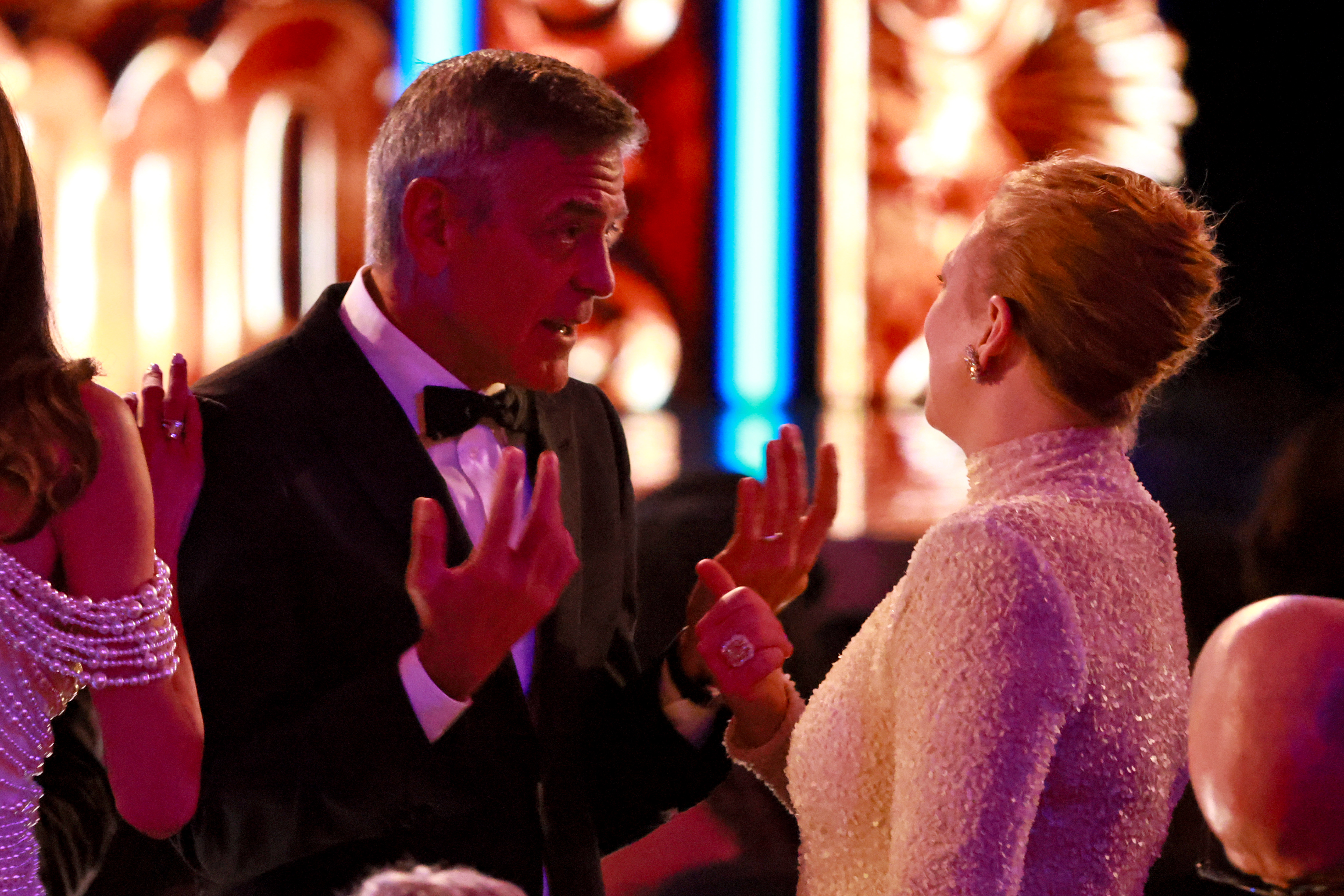 With expressive hand gestures, George Clooney chats with Sarah Snook mid-show as Amal Clooney stands beside him. The animated moment unfolds in the warmly lit theater.
