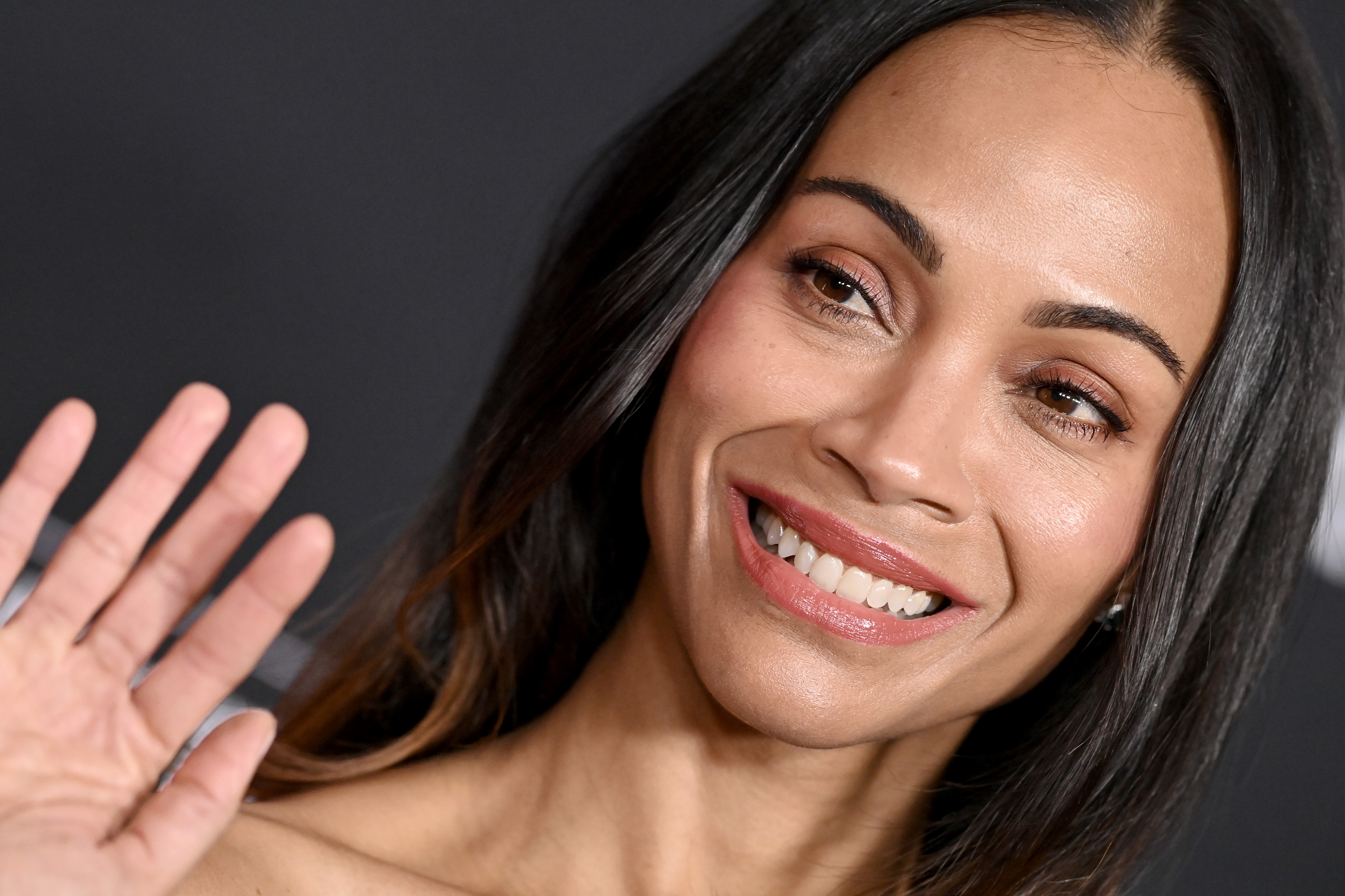Zoe Saldaña flashes a radiant smile and waves to fans, showcasing her natural beauty with glowing skin, soft makeup, and sleek, straight hair at the "Avatar: Fire and Ash" premiere.