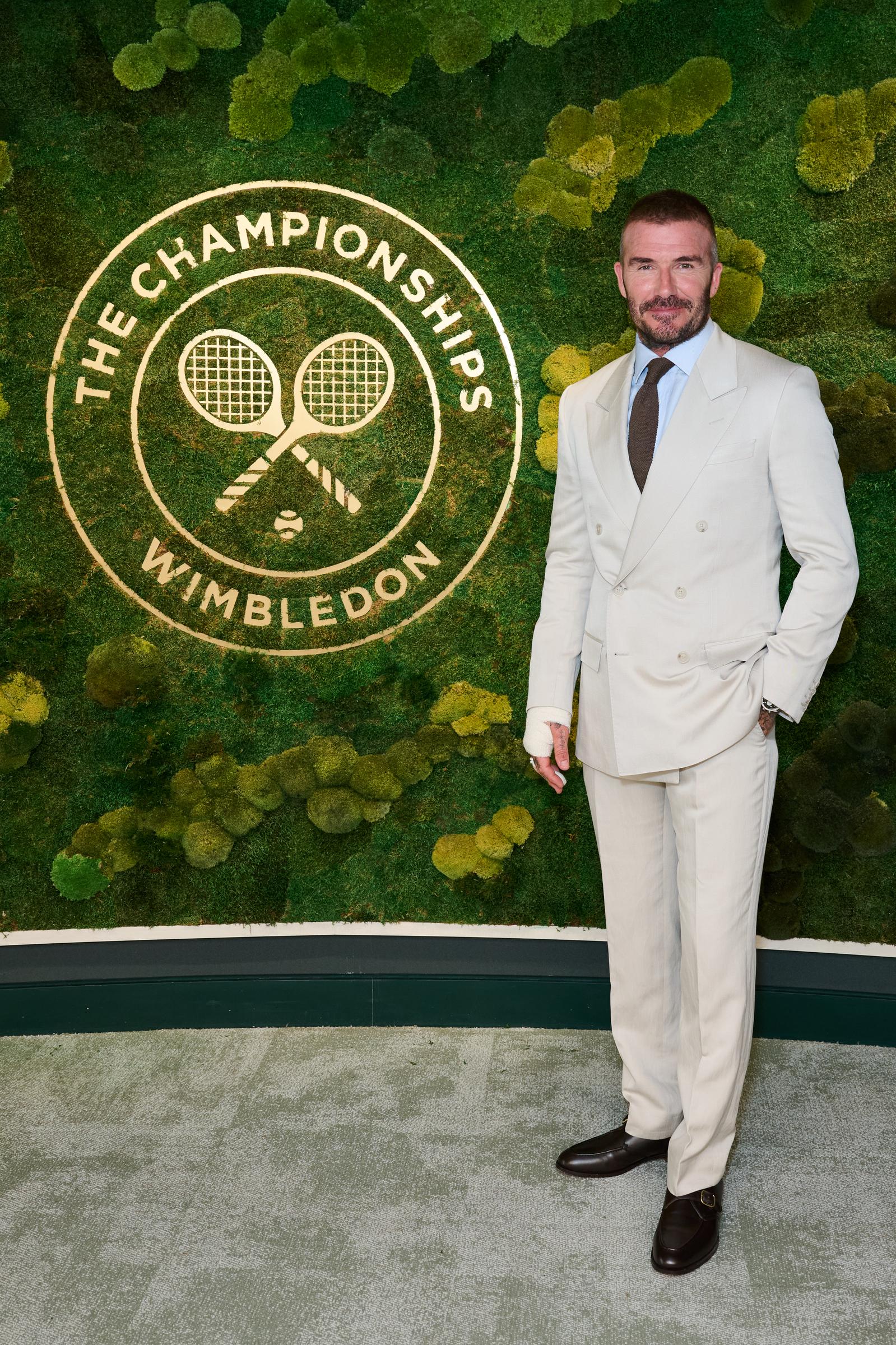 David Beckham pauses for a solo portrait at Wimbledon, standing confidently in front of the iconic moss wall at the AELTC Parkside Suite. His tailored cream suit and bandaged hand subtly reflect his enduring poise and style.