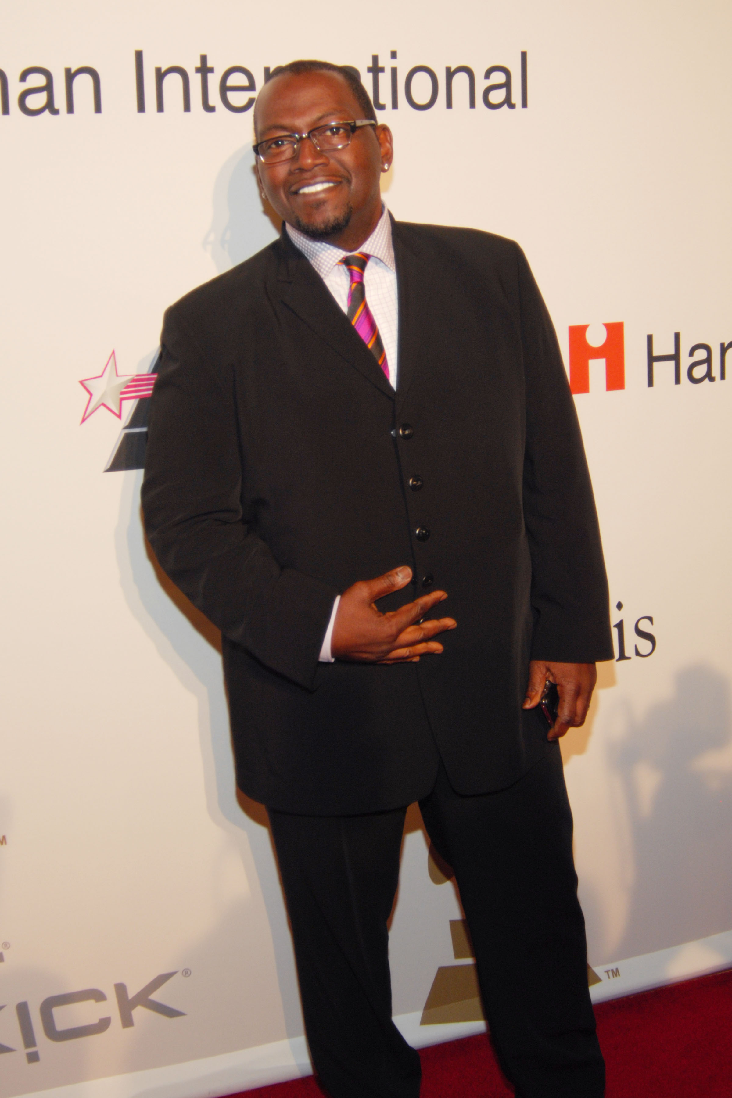 Dressed in a tailored black suit with a colorful striped tie, Randy Jackson smiles confidently while posing on the red carpet.