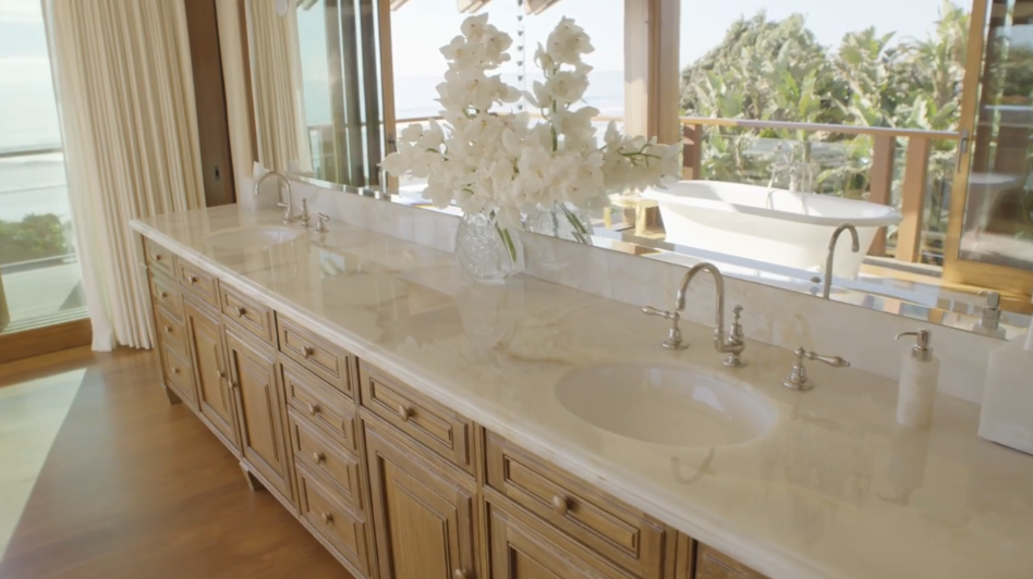 A long double vanity featuring light polished marble countertops and custom wood cabinetry anchors this spacious bathroom area. A full-length mirror reflects lush greenery and a white freestanding tub from the adjacent room, while a large vase of white orchids creates a refined focal point. | Source: YouTube/Architectural Digest