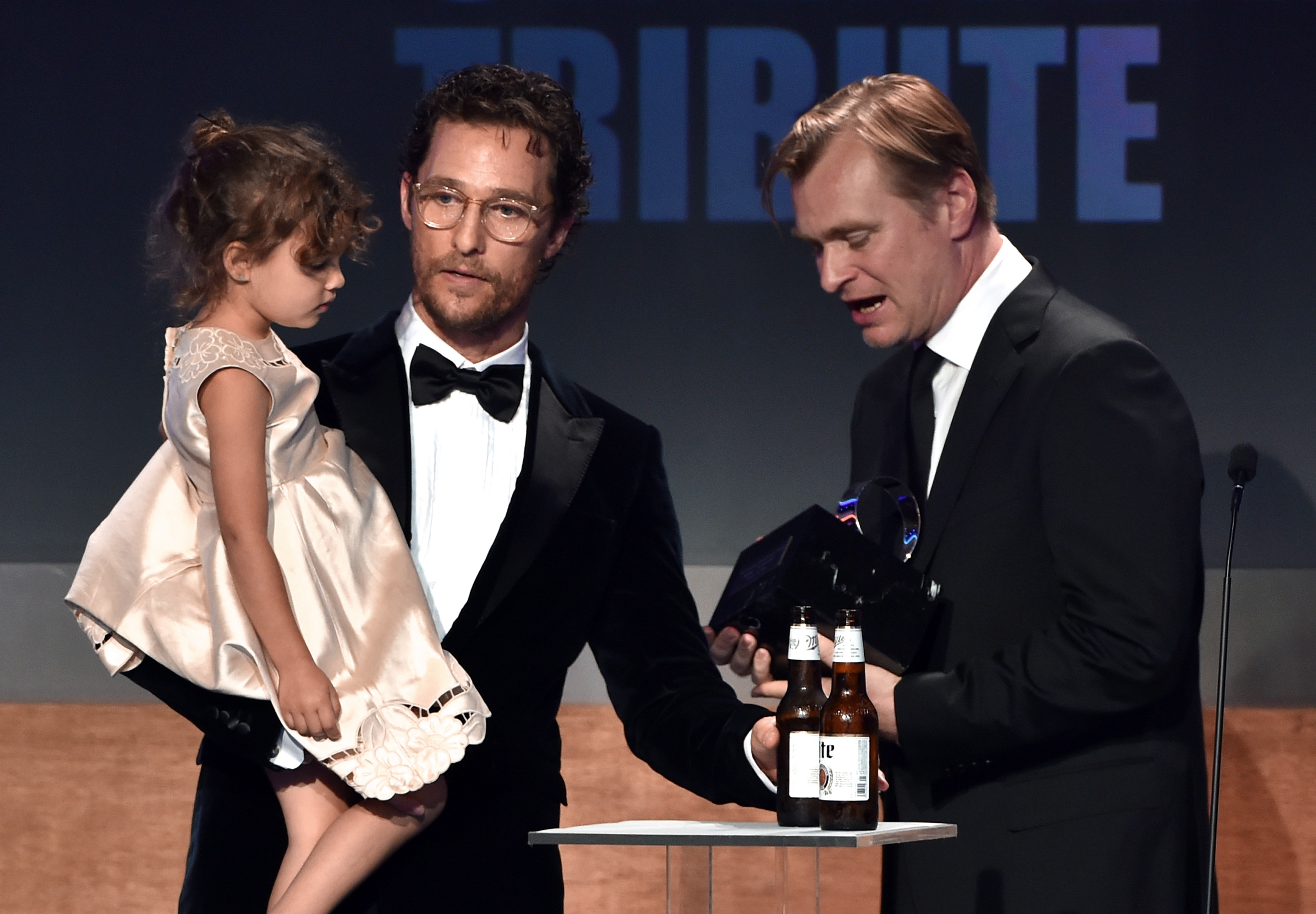 Vida Alves McConaughey is held on Matthew McConaughey's arm while onstage. She wears a light dress and looks downward as he stands in a tuxedo. Christopher Nolan stands beside them holding an award and speaking.