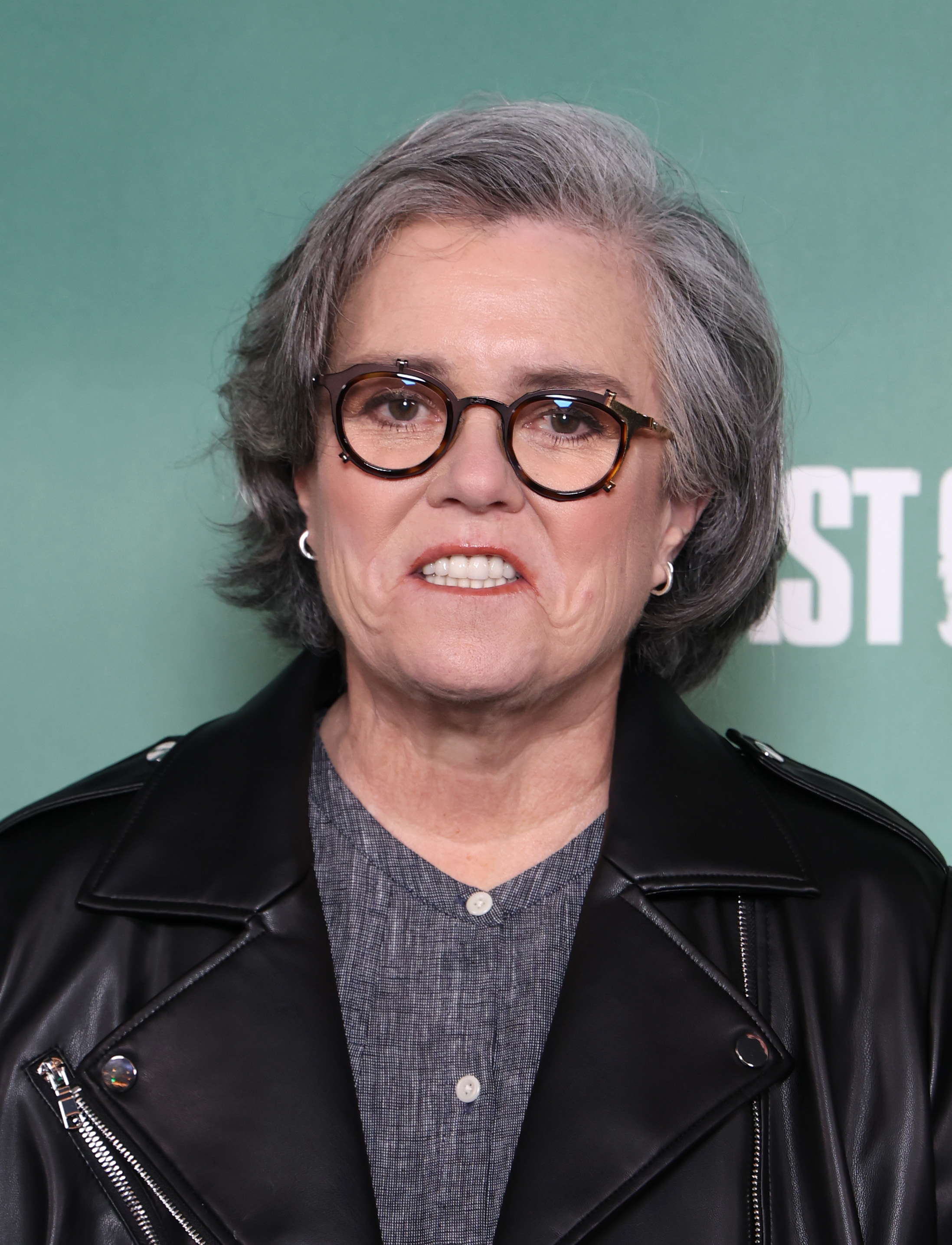 Rosie O'Donnell attends the London premiere of "The Last of Us" Season 2, posing against a green backdrop in a black leather jacket and gray top with round glasses.