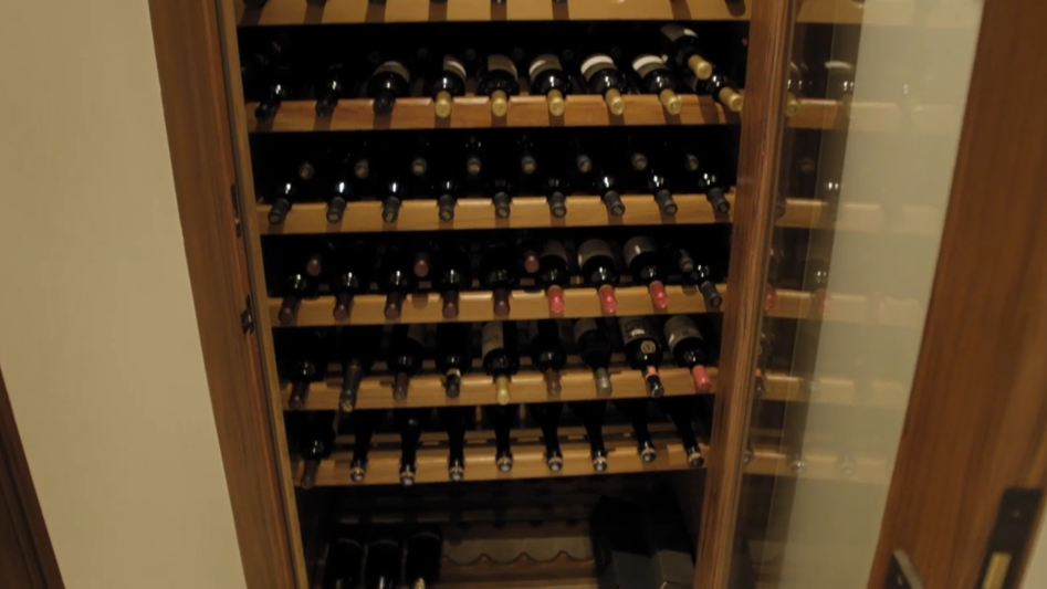 A temperature-controlled custom wine storage cabinet built into rich wood framing houses numerous bottles arranged neatly across multi-tier wooden racks. Clear glass door panels allow for seamless viewing of the extensive collection while maintaining optimal storage conditions. | Source: YouTube/Architectural Digest