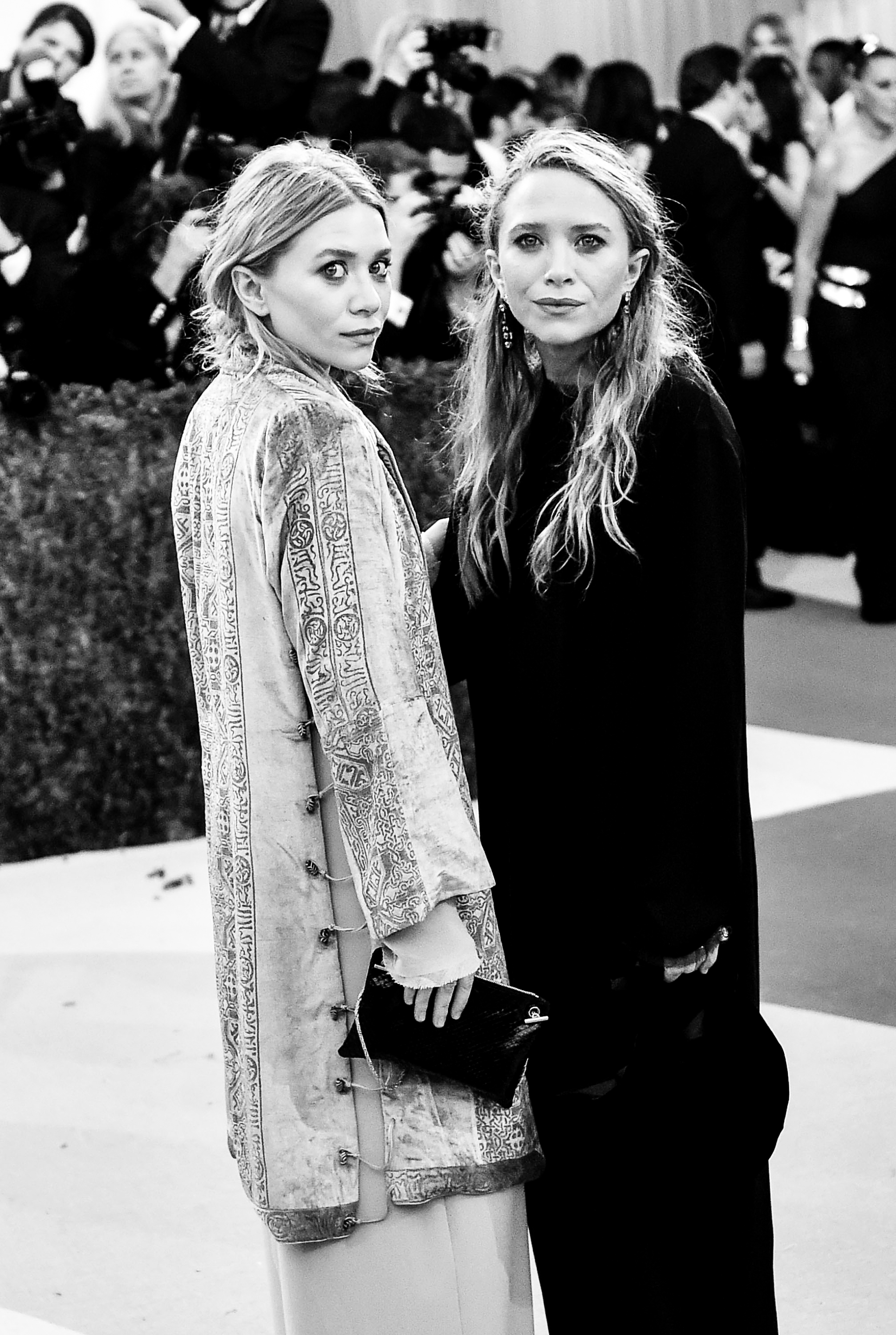 Mary-Kate and Ashley Olsen stand side by side at the Met Gala, turning slightly toward the camera. The black-and-white shot highlights their layered looks and calm, composed expressions.
