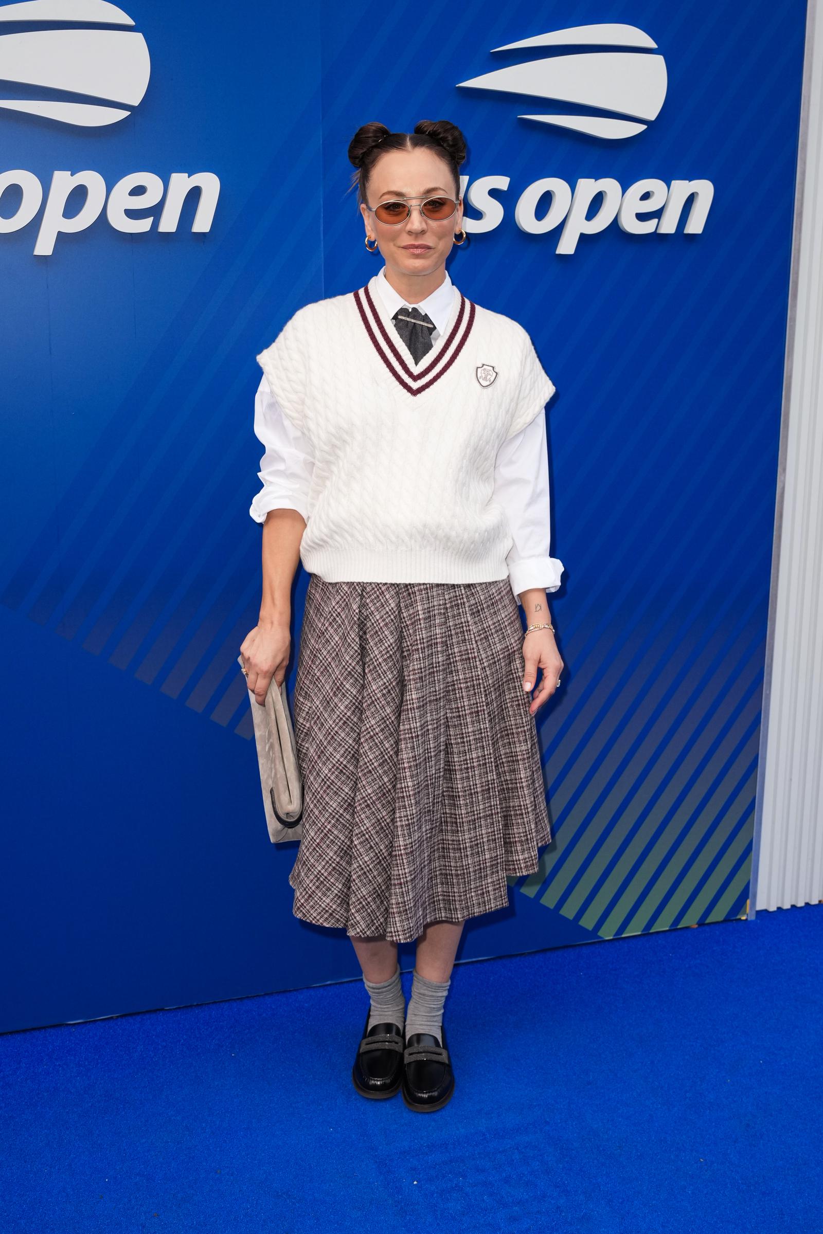 Kaley Cuoco attends the US Open in a preppy, school-inspired look featuring a cable-knit tennis vest over a white button-up shirt with a plaid midi skirt. She finishes the outfit with black loafers, gray socks, round sunglasses, and a playful double-bun hairstyle.