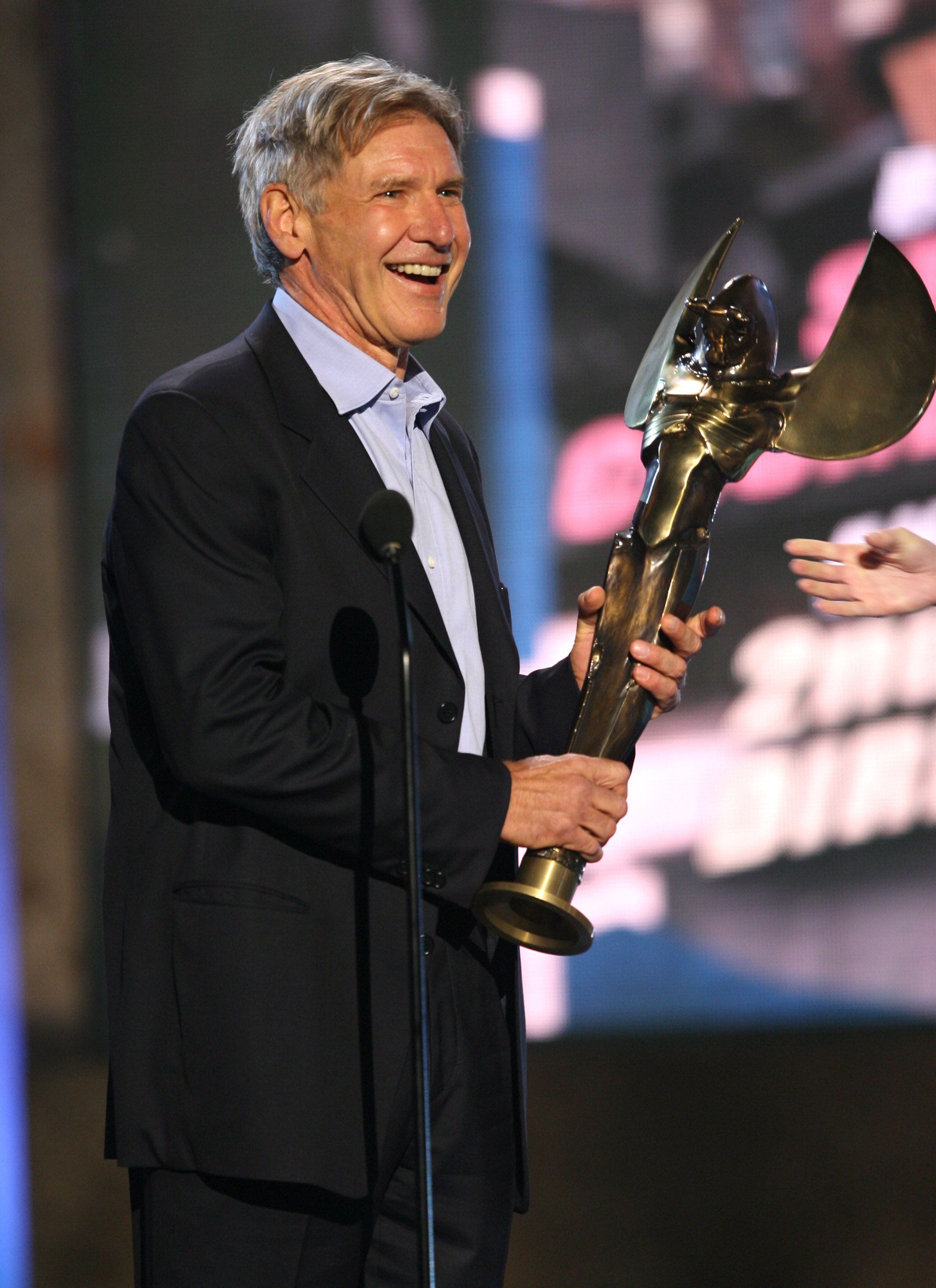 Clutching a winged golden trophy, a beaming Harrison Ford shares a laugh on stage at Paramount Studios during the Taurus World Stunt Awards. The actor attended the event in a dark suit jacket worn over an unbuttoned light blue dress shirt. | Source: Getty Images