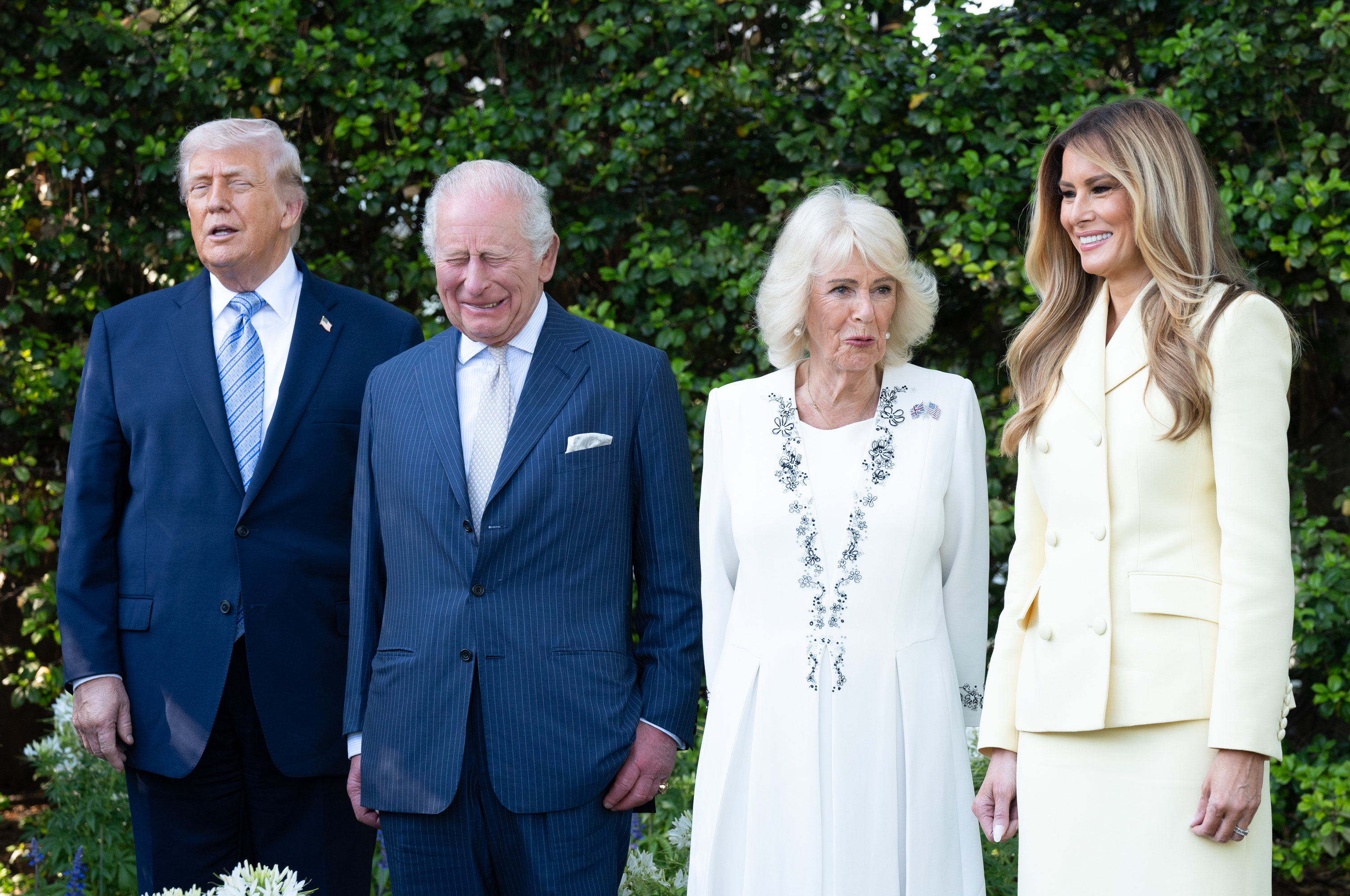U.S. President Donald Trump, King Charles III, Queen Camilla, and First Lady Melania Trump stood in a line for a posed photograph against a backdrop of dense greenery, capturing a composed yet slightly candid moment during the afternoon tea on the White House grounds. The arrangement highlighted the visual contrast between the couples, with formal attire and expressions underscoring the ceremonial weight of the occasion.