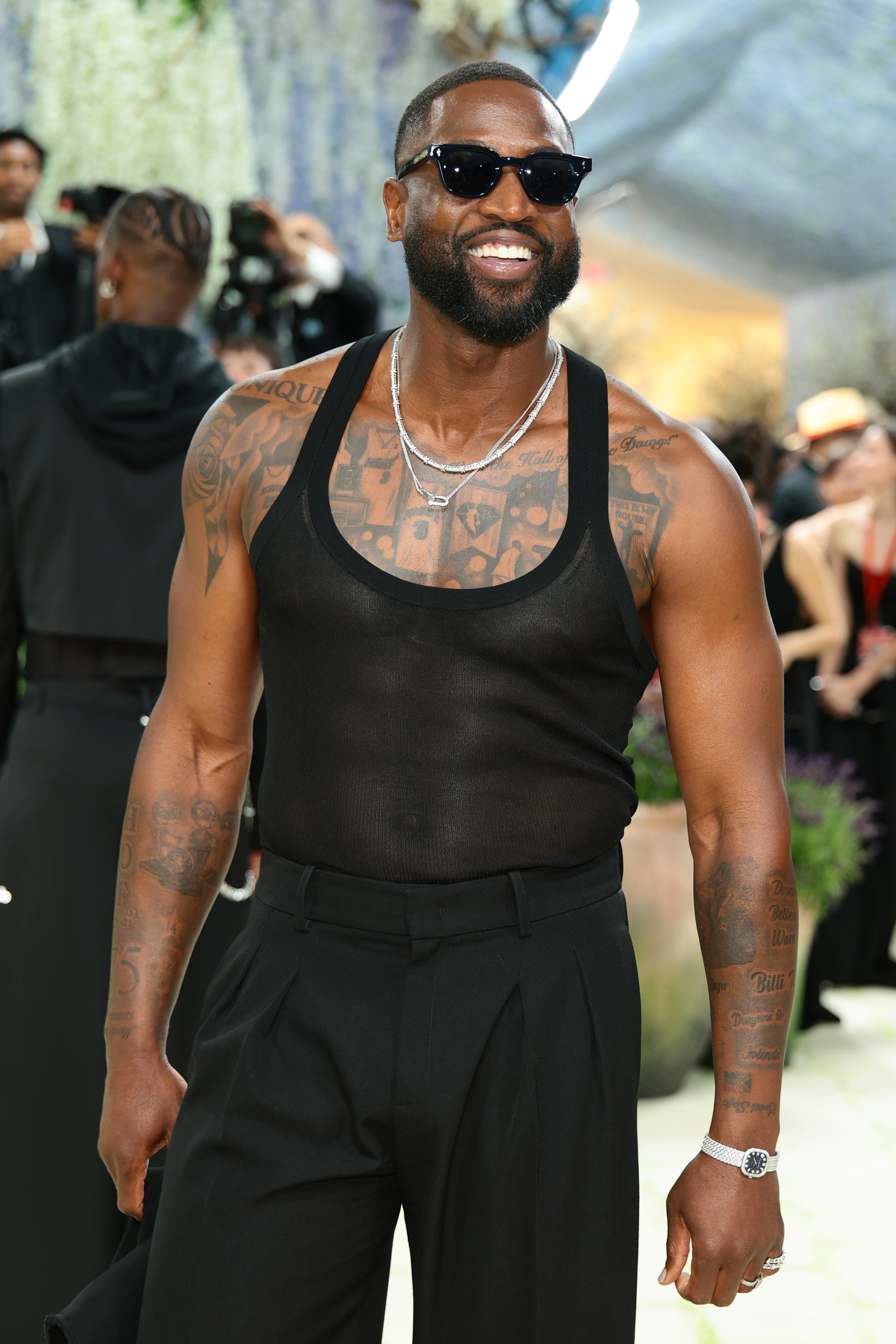 Dwyane Wade steps out in a sheer black tank paired with tailored trousers, his layered chains and dark sunglasses adding a sleek, modern edge. His confident smile and tattooed chest on display turn the minimalist look into a bold, statement-making moment.