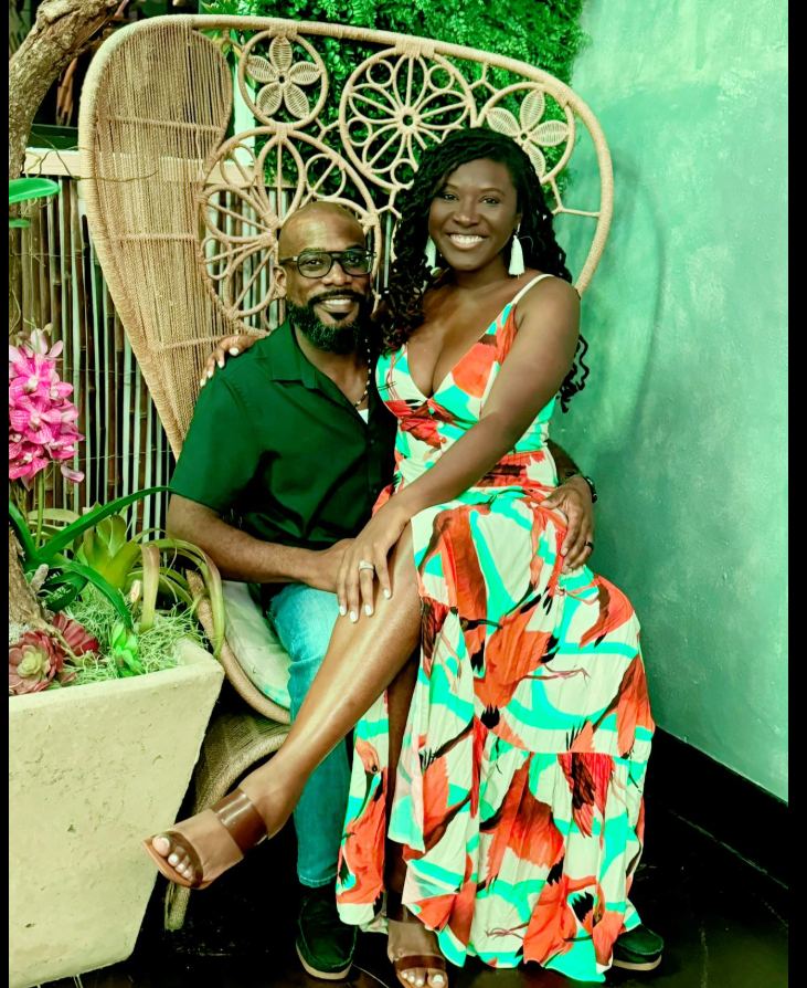 Seated together in a cozy, garden-like setting, Nancy Metayer Bowen smiles brightly in a flowing, colorful maxi dress while her husband, Stephen Bowen, sits beside her with one arm around her waist, the pair framed by a decorative wicker chair and lush greenery. | Source: Facebook/Nancy Metayer Bowen