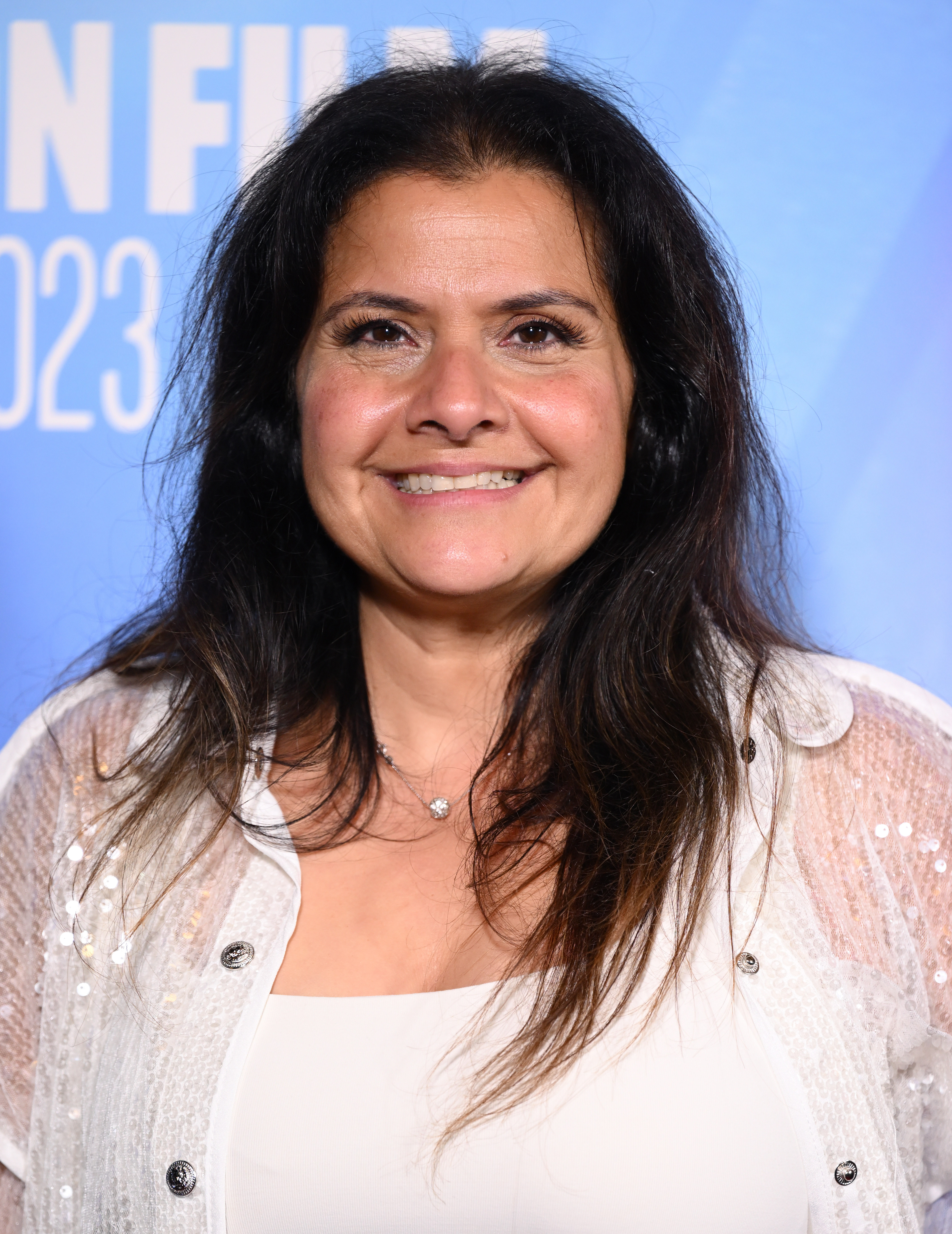 A radiant close-up of Wadia at the BFI London Film Festival premiere of "Bonus Track" at Vue West End in October 2023, her dark hair worn loose and unstyled around her face. She wears a cream white top layered under a sparkly white embellished cardigan with silver button detailing, and a delicate pendant necklace, her complexion visibly clearer and her face noticeably slimmer than in earlier years.