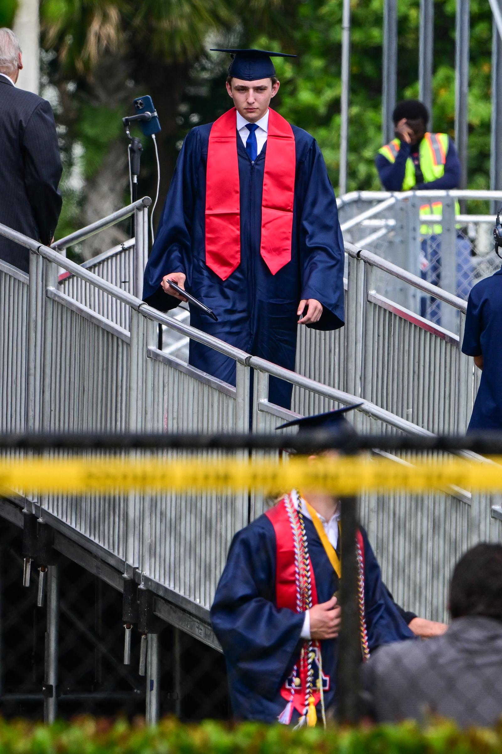 Barron Trump walks down a ramp in his cap and gown during his graduation ceremony at Oxbridge Academy in Palm Beach, Florida. Dressed in navy robes with a bold red stole, he holds his diploma as he heads toward the stage.