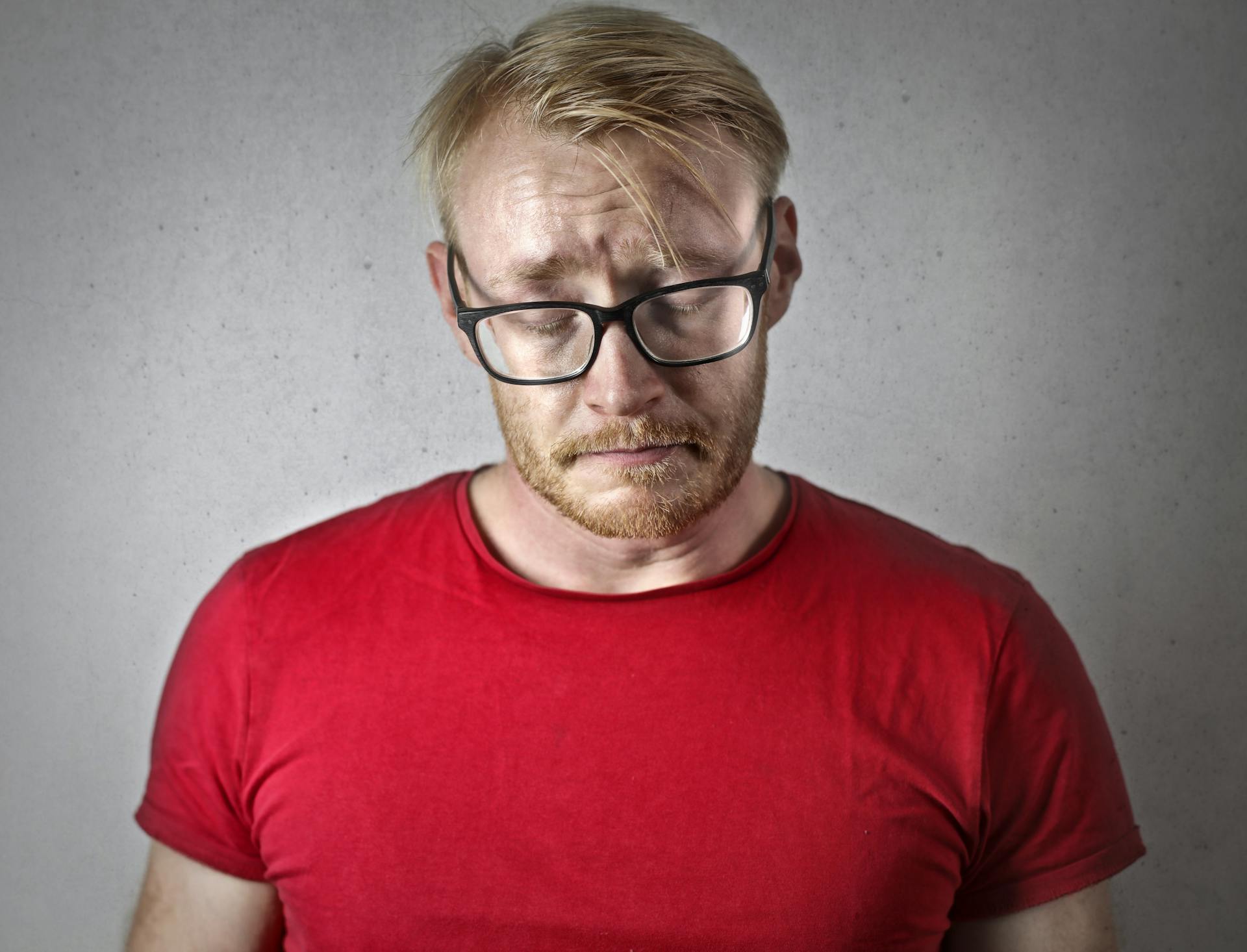 Sad man in red T-shirt and black framed glasses closing his eyes | Source: Pexels