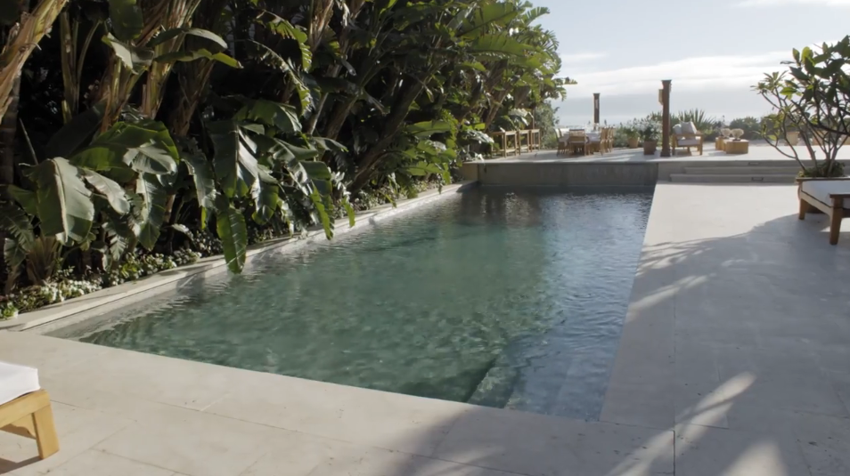 Surrounded by dense banana palms and light-toned travertine paving, the rectangular saltwater swimming pool stretches peacefully along the side of the estate. Sun loungers sit on the surrounding terrace, while an elevated dining deck and open coastal views complete the resort-style outdoor area. | Source: YouTube/Architectural Digest