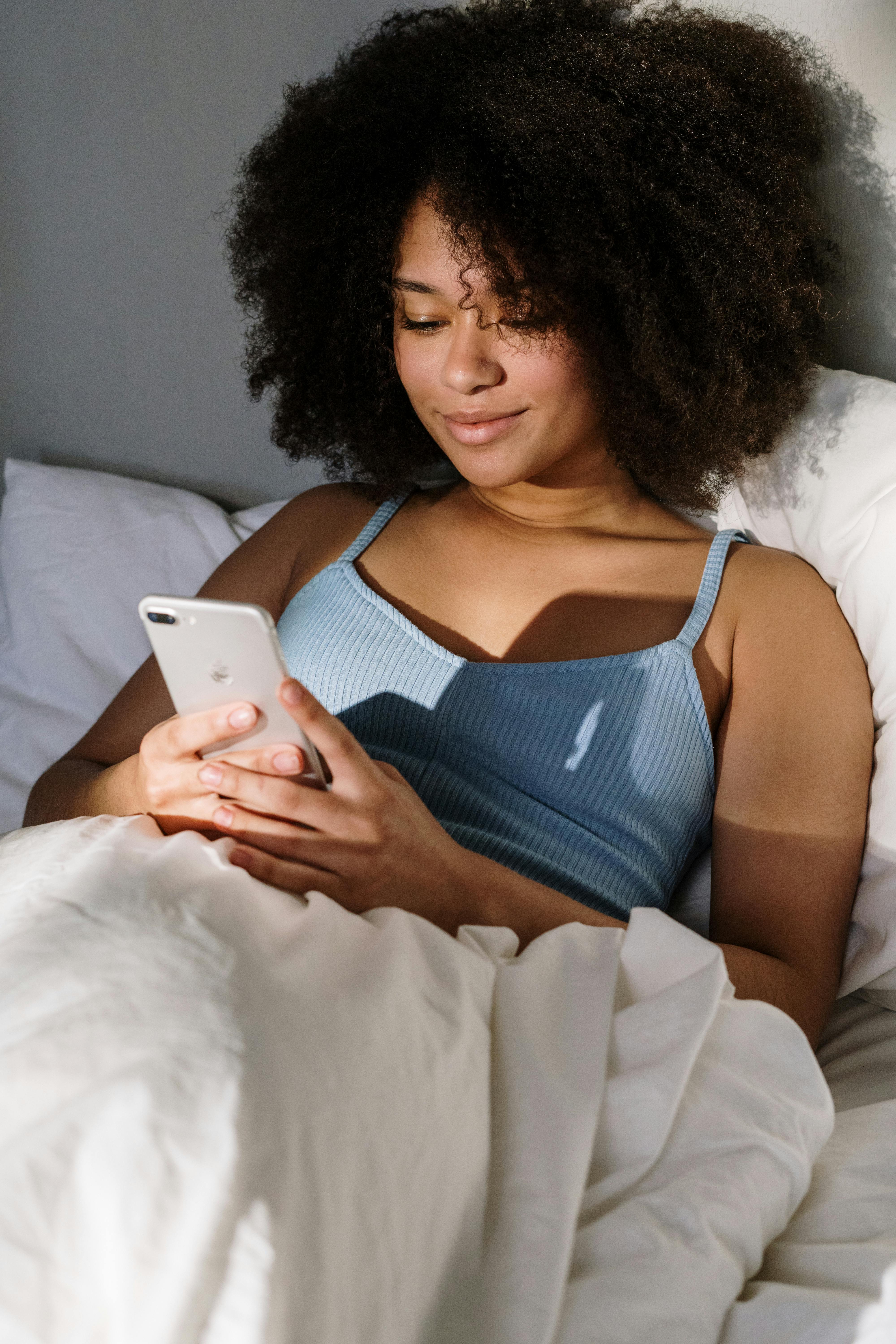 A woman smiling while looking at her phone in bed | Source: Pexels