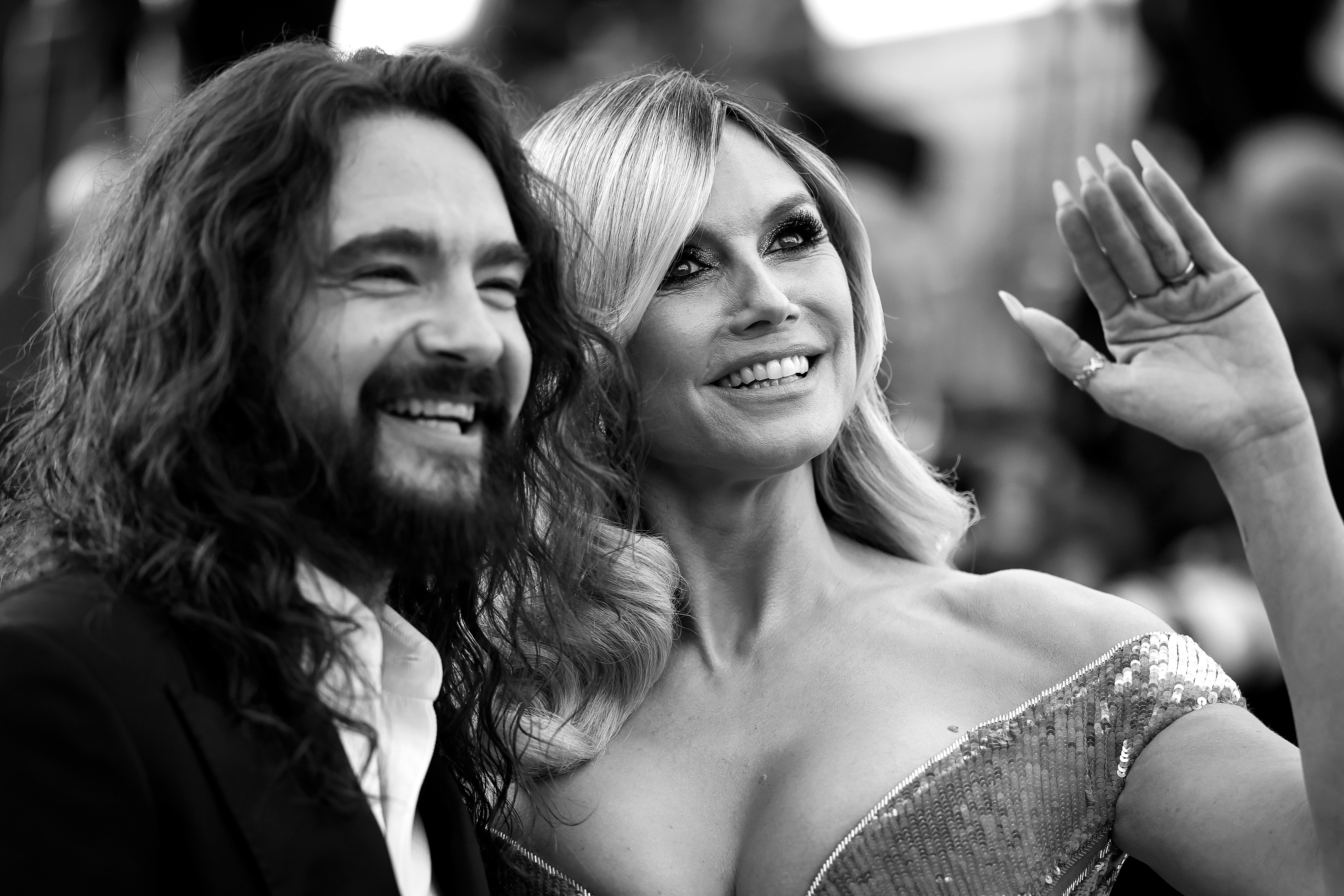 Kaulitz and Klum's faces are both clearly visible as they look toward the camera. He is beaming with pride, his wide smile reflecting the joy of supporting his wife. Beside him, Klum raises her hand in a graceful wave.
