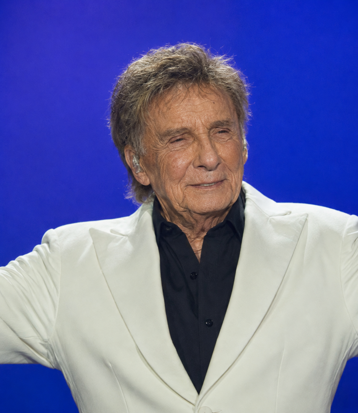 The open-armed pose and white jacket preserve Manilow's familiar showman image. The rendering changes the face while leaving the performance style intact, making the contrast more noticeable. | Source: ChatGPT