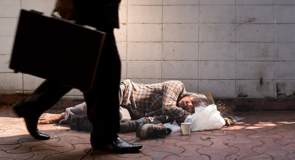 Hombre sin hogar durmiendo en el suelo mientras una persona camina frente a él. | Foto: Shutterstock