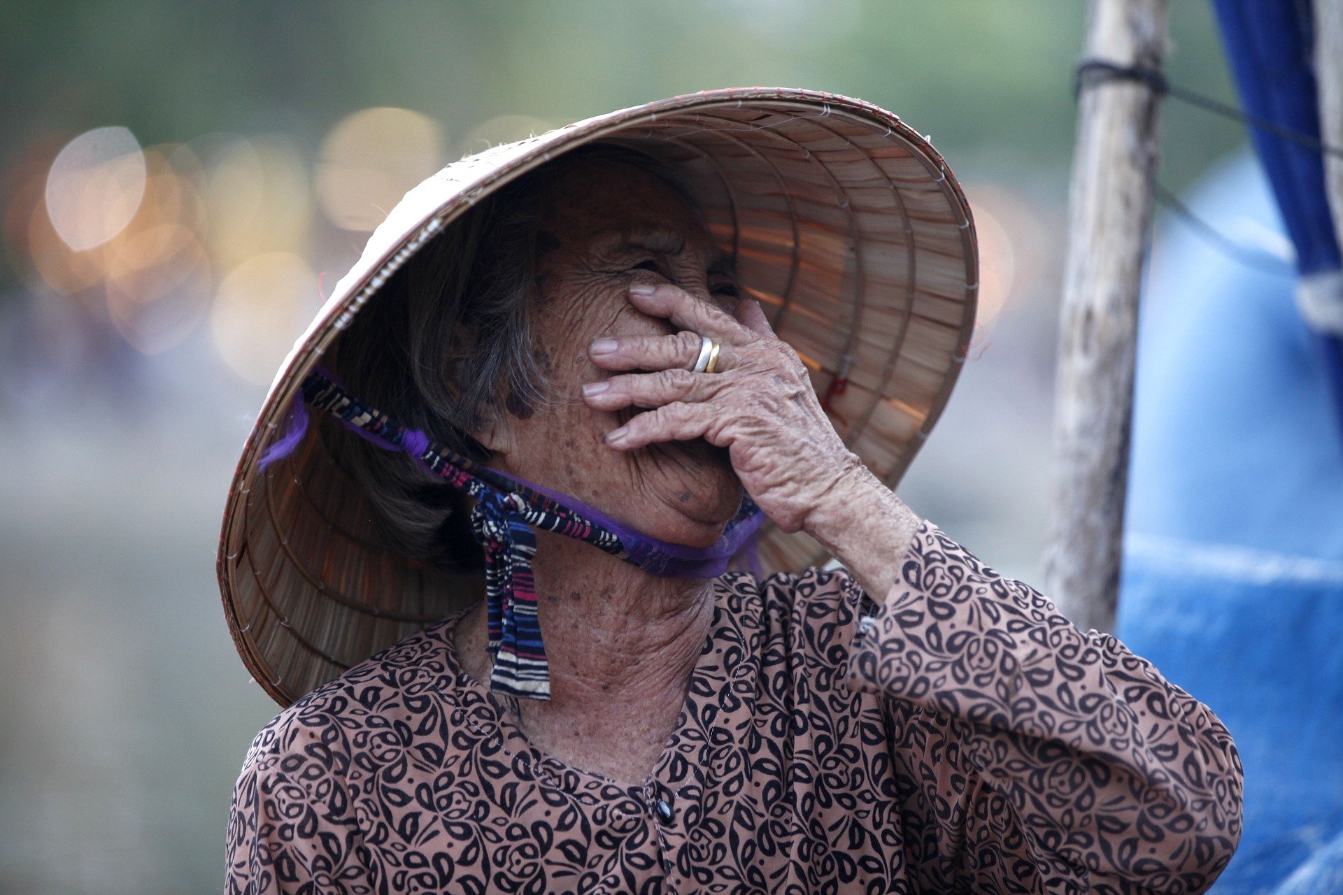 An old woman wearing a hat, covering her mouth while laughing | Photo: Pixabay/Thangphan