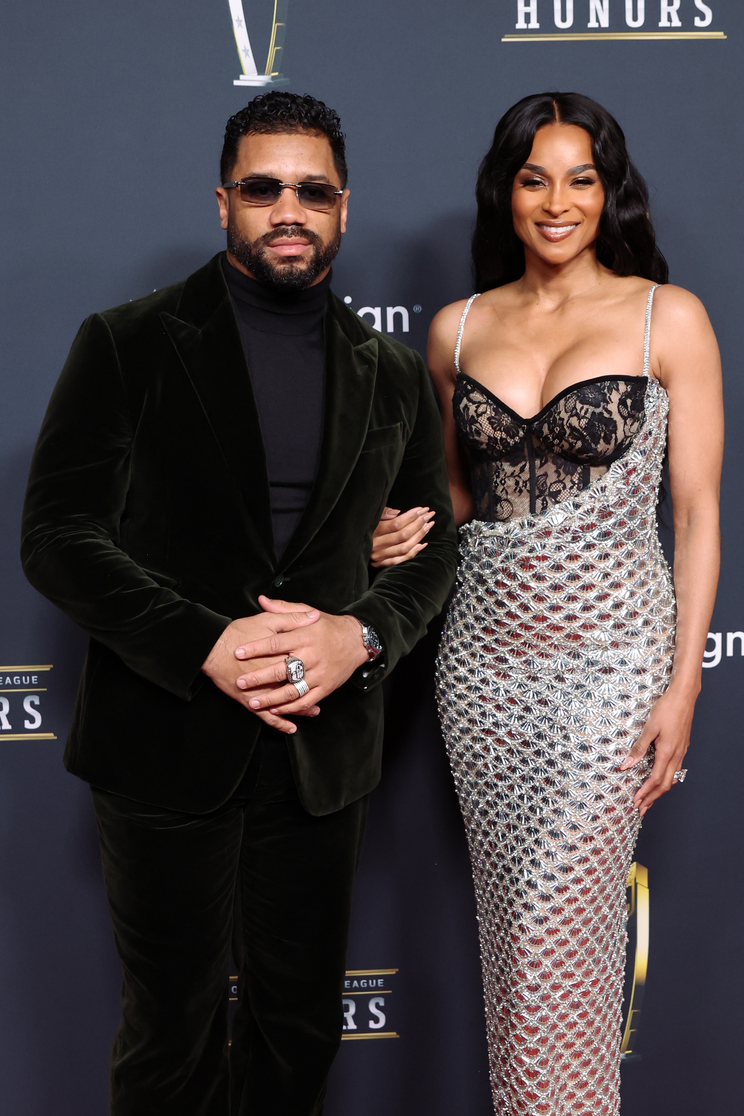 Russell Wilson stands beside Ciara in a dark velvet suit, while Ciara shines in a silver gown with a corset-style bodice, the couple posing confidently as one of the evening's most recognizable power pairs.