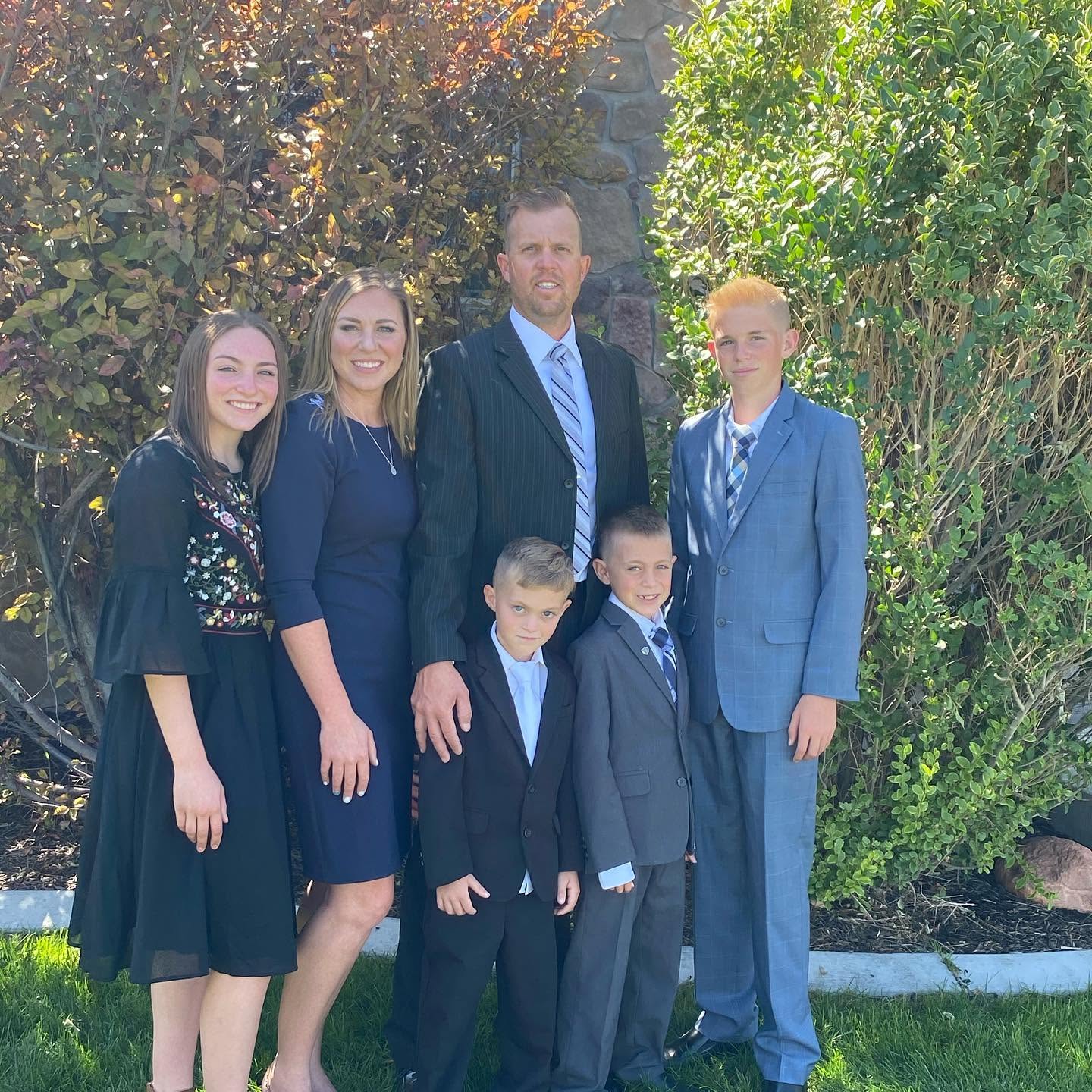 The entire Long family poses formally outdoors against a backdrop of green foliage and a stone wall. Katrina stands elegant in navy, Lydia in a detailed black dress, and Captain Spencer looks sharp alongside his three sons, Reid, Thayne, and Gage, all dressed in their sharpest suits. | Source: Facebook/katrina.g.long